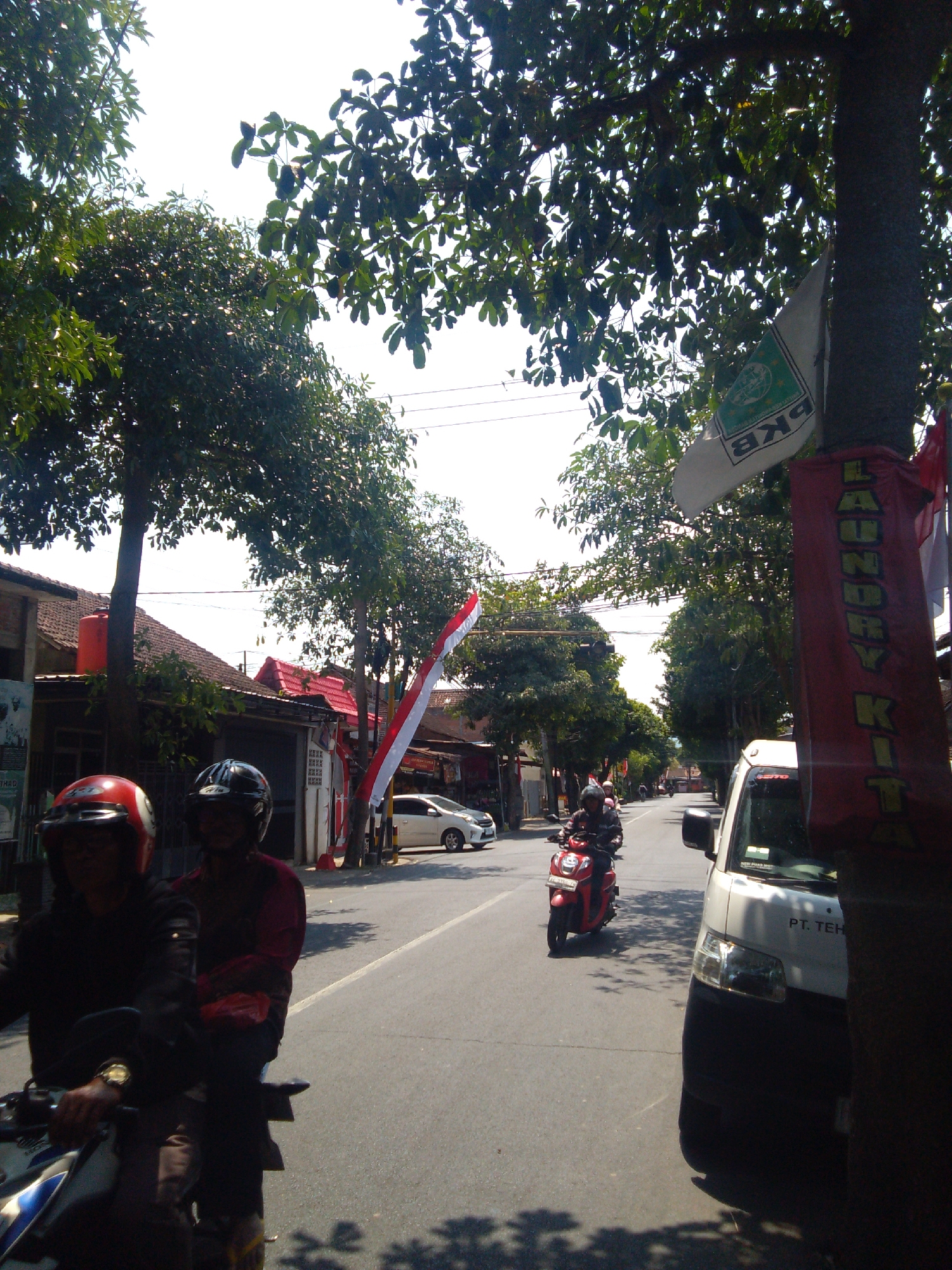 A sunny, daytime scene depicts a street in Trenggalek, Indonesia, lined with trees and low-rise buildings. In the foreground, two individuals wearing helmets are on a motorbike. The rider in the front wears a red and white helmet, while the passenger behind wears a black helmet and a maroon shirt. A red bag is visible between them.
Further down the street, a red motorbike with a rider wearing a black helmet is traveling away from the camera. A white van is parked on the right side of the road. Red and white Indonesian flags are visible on poles along the street, as is a banner with the word "LAUNDRY KIT" written vertically in yellow letters. The road is paved, and shadows from trees are cast on the pavement. Buildings on the left appear to be shops or homes with tiled roofs. The sky is clear and bright.