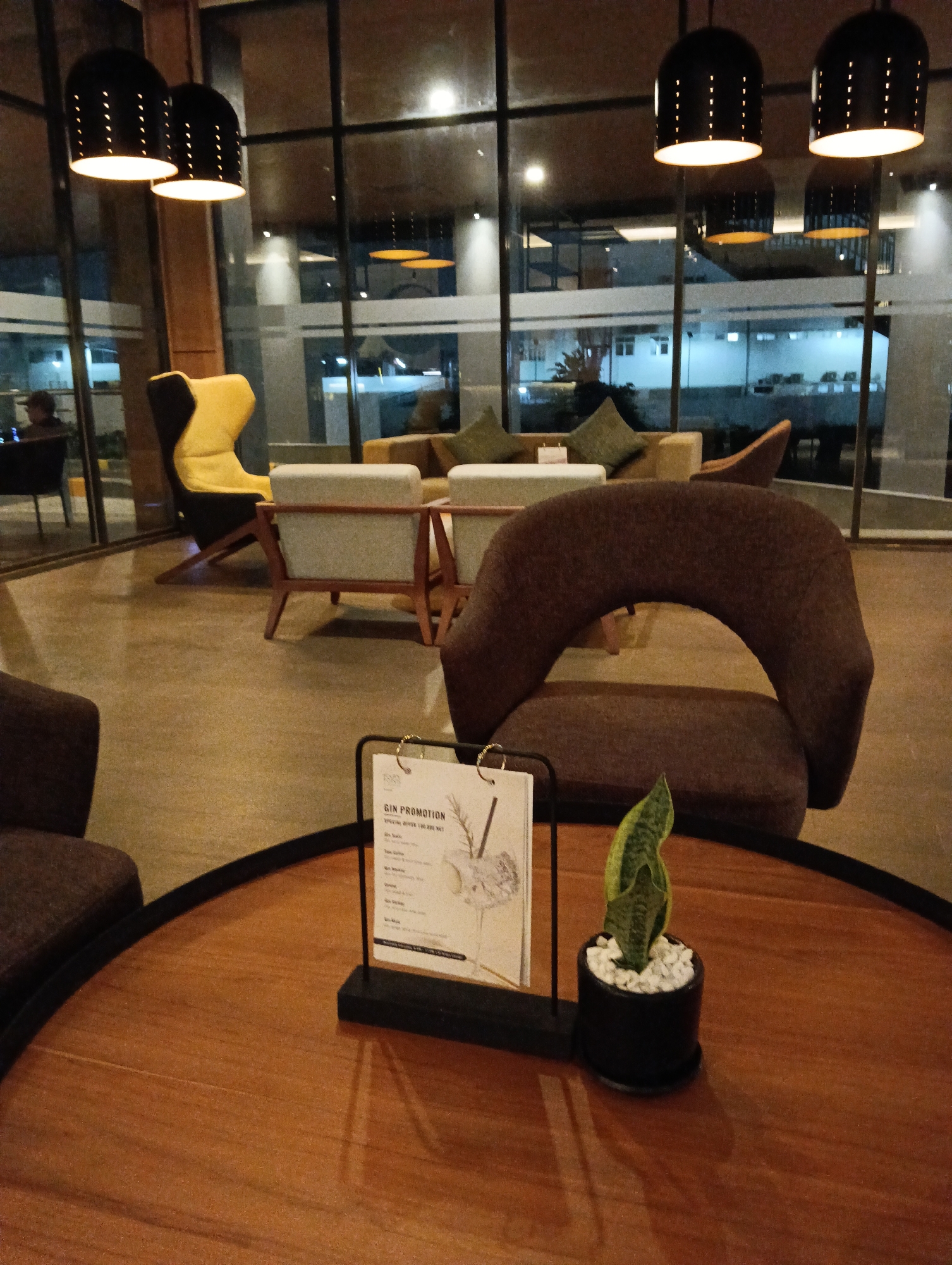 A low-angle shot captures a modern lounge area with large windows overlooking an outdoor scene at dusk. The interior features a round, polished wooden table in the foreground, adorned with a small potted snake plant and a menu holder. Several seating arrangements are visible, including a distinctive brown upholstered chair with a curved back, two grey upholstered armchairs with wooden frames, and a bright yellow armchair. In the background, through the expansive windows, lights from buildings and streetlights illuminate the exterior. The lighting within the lounge is warm, emanating from pendant lamps suspended from the ceiling and ambient lighting from the windows. A person can be seen in the far left background, seated at a table. The overall impression is of a sophisticated and contemporary public space, likely a hotel lobby or a high-end cafe, situated in Sungai Raya, Indonesia.