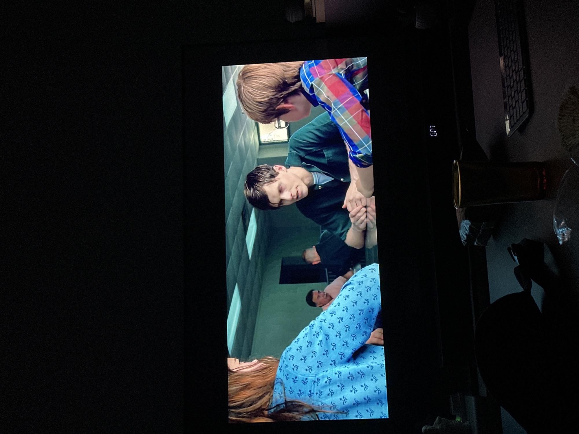 The image is a photograph of a television screen displaying a scene from a movie or television show. The scene depicts a hospital setting. A woman with long brown hair lies in a hospital bed, wearing a light blue patterned hospital gown. Two men are standing over her. One man is wearing a dark green shirt. The other man is wearing a plaid shirt and glasses. Other patients are visible in the background of the shot. The location in the city of Lovech, Bulgaria is not visible in the image. Other objects such as a keyboard, a can, and a water bottle are present in the room where the picture was taken. The room where the picture was taken is dark.