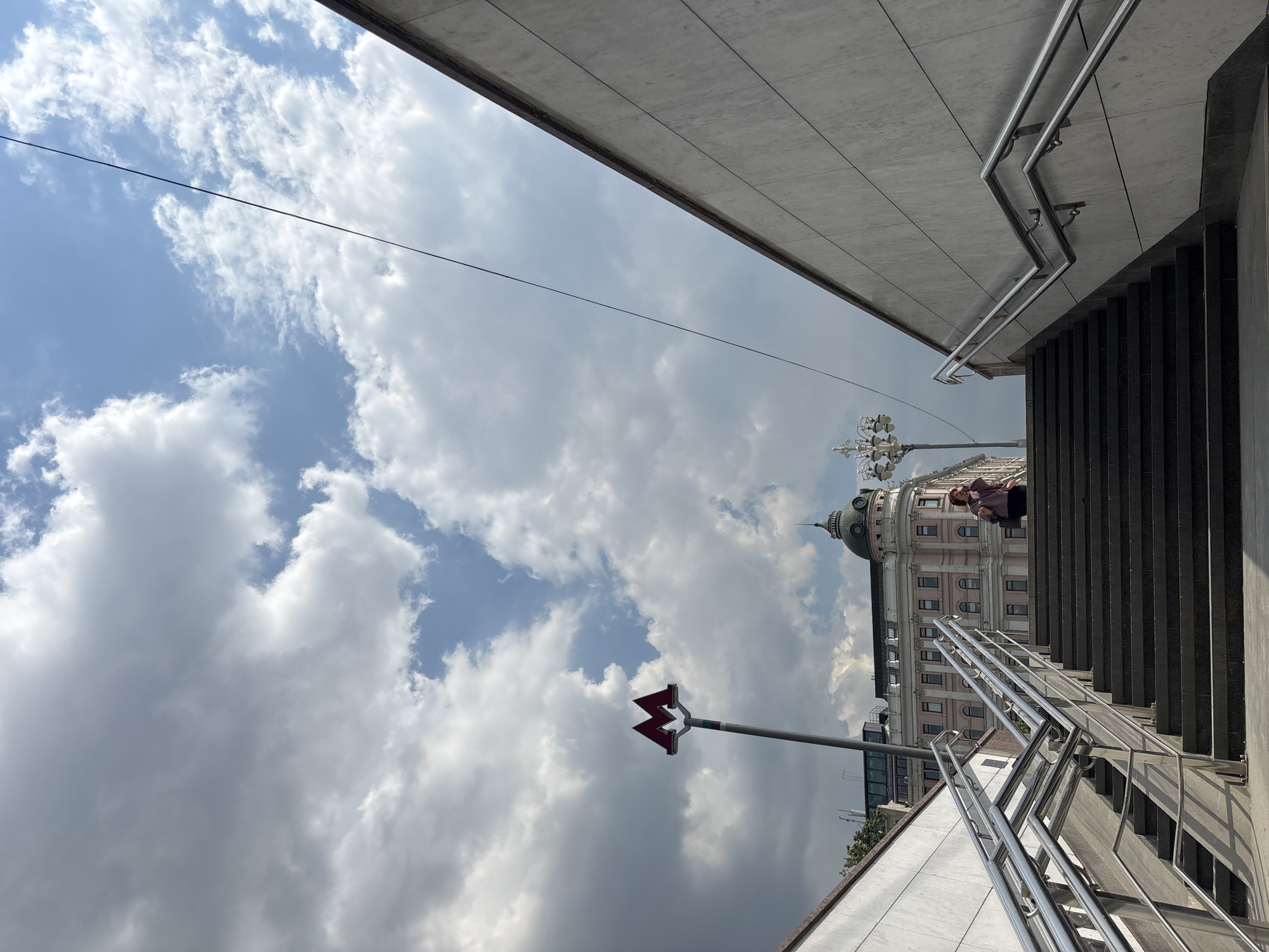 The image depicts a view of a Moscow metro entrance on a cloudy day. In the foreground, a wide staircase with metallic railings leads upwards. To the left, a segment of a white stone wall is visible. To the right, a similar white stone wall angles upwards. A person is positioned on the staircase, wearing a dark outfit and carrying a bag. In the background, a pink building with a green dome stands out, topped with a spire. A classic streetlamp is situated in front of the building. A red "M" sign, indicating a metro station, is mounted on a pole to the left of the pink building. The sky is filled with dramatic clouds, with patches of blue visible. A thin black wire stretches diagonally across the upper portion of the frame.