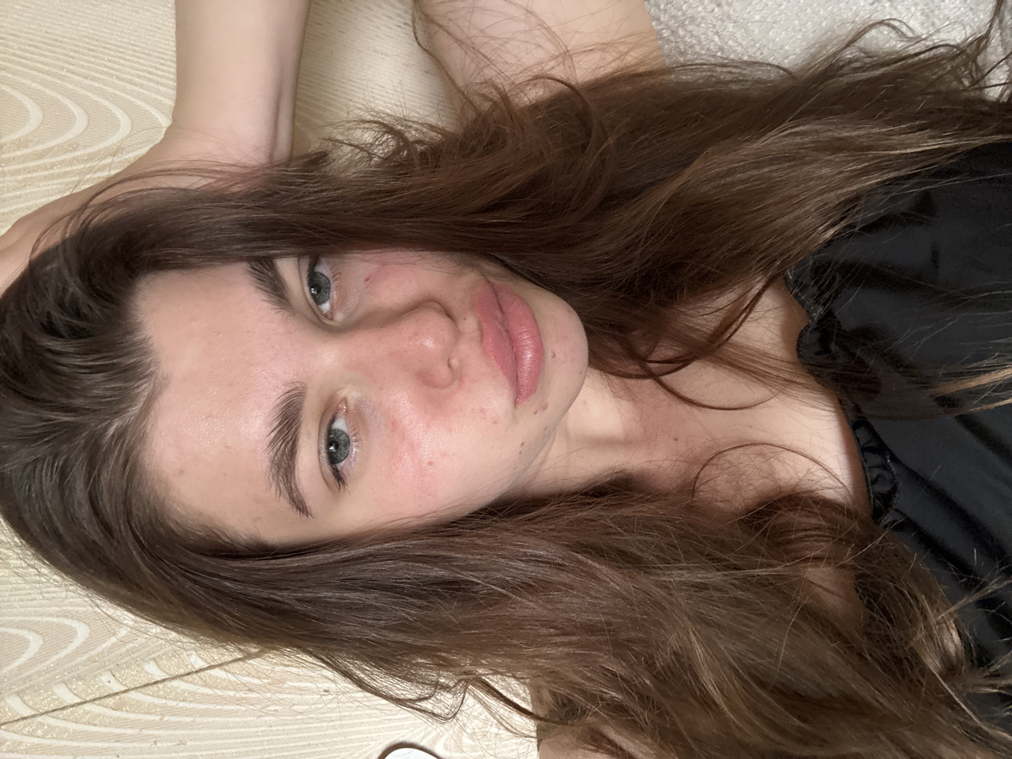 A young woman with light skin, light eyes, and long brown hair is lying down, looking directly at the camera. She is wearing a black top. The background is a beige surface with a subtle patterned texture. The location is not discernible from the image provided.