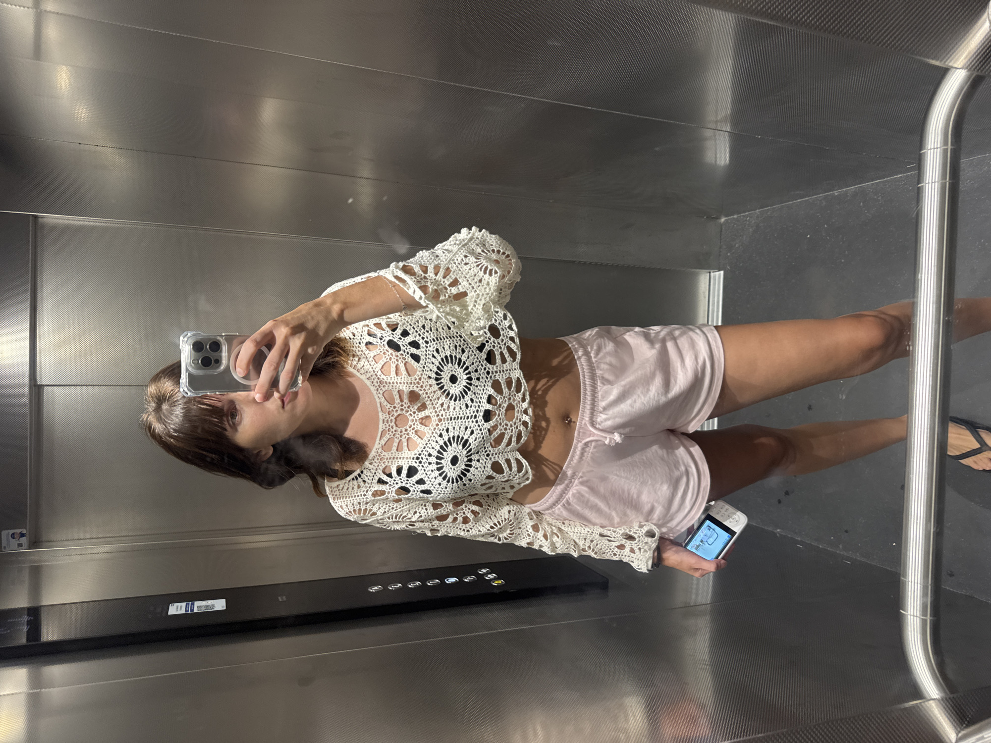 A woman is standing in an elevator, taking a selfie with her phone. She is wearing a white crochet crop top, pink shorts, and black flip-flops. She has brown hair and is holding a phone in her left hand. The elevator has silver walls and black buttons. The floor is black. The elevator appears to be in a building located in Isola delle Femmine, Italy, based on the presence of Italian text on a sticker on the elevator wall.