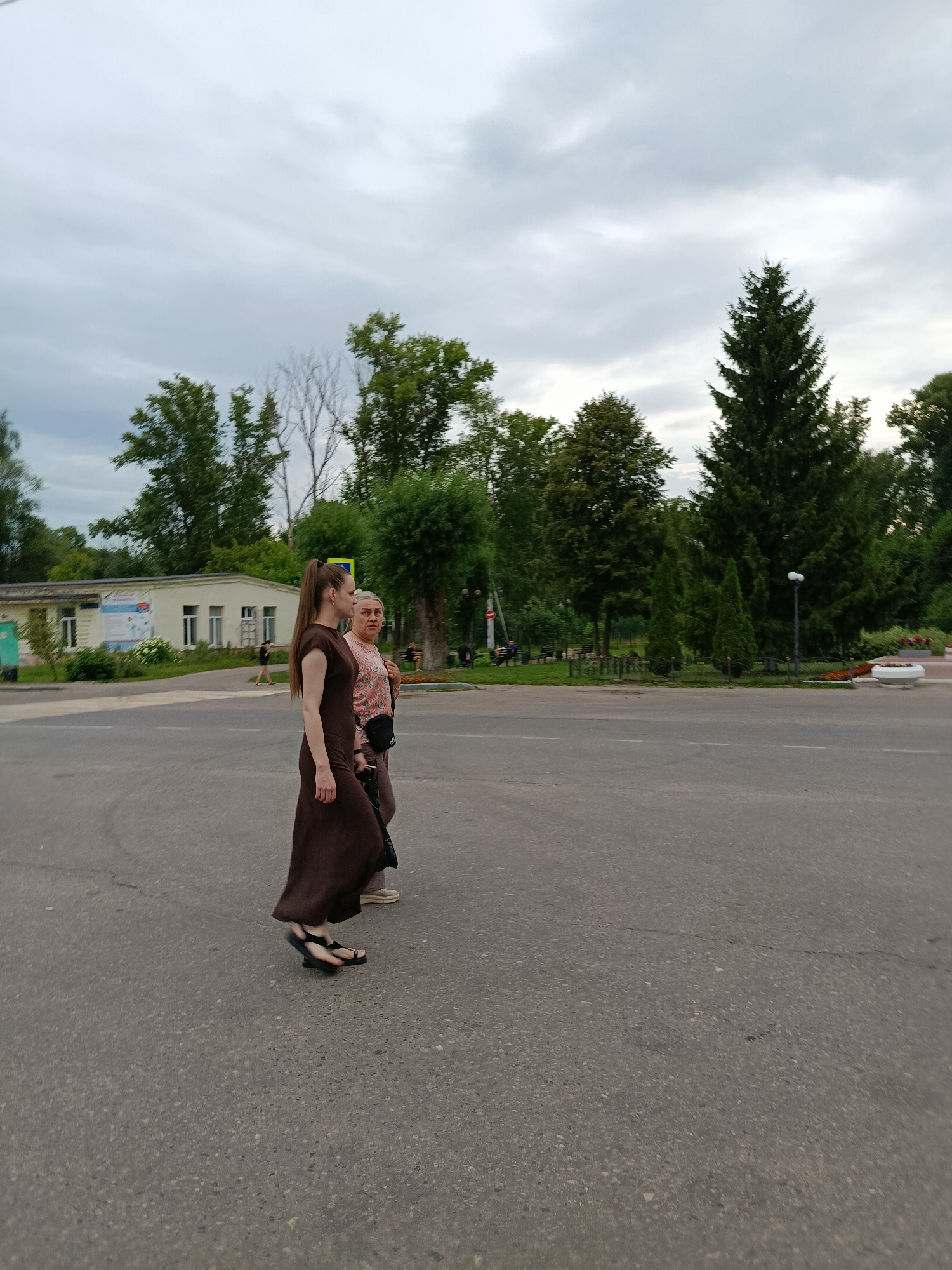 Two women are walking across a street in Verea, Russia. The woman in the foreground is wearing a long, dark brown dress and black sandals. She has her hair pulled back in a ponytail. The woman behind her is wearing a light-colored patterned shirt and dark pants, with a black bag slung over her shoulder. She appears to be looking at something to her right.
The scene is an outdoor setting with trees and a building in the background. The sky is overcast, suggesting cloudy weather. The time of day appears to be late afternoon or early evening, as the light is soft. The street is paved, and there are white lines marking a crosswalk. In the distance, there are more trees and some park benches. There is also a lamppost visible. The overall mood seems to be casual, with the two women engaged in conversation or simply walking together. There is no visible text in the image.