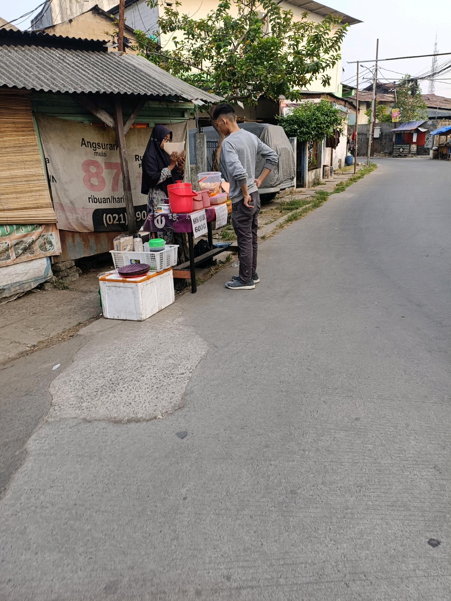 A street vendor, a woman wearing a black hijab and patterned dress, is interacting with a male customer, who is wearing a grey long-sleeved shirt and dark pants. The vendor is handing a small paper-wrapped item to the customer. They are standing at a stall set up on the side of a paved road in Tangerang, Indonesia. The stall is covered by a corrugated metal roof and displays various food items in containers and baskets, along with a red bucket and a white styrofoam cooler. A banner behind the vendor reads "Angsuran mulai 87 ribuan/hari" (Installment starts from 87 thousand per day) and displays a phone number, "(021) 90xxx." The environment appears to be an urban setting with buildings and trees visible in the background. The road is slightly sloped, and the pavement is cracked and uneven in places. There are other stalls and structures further down the road.
