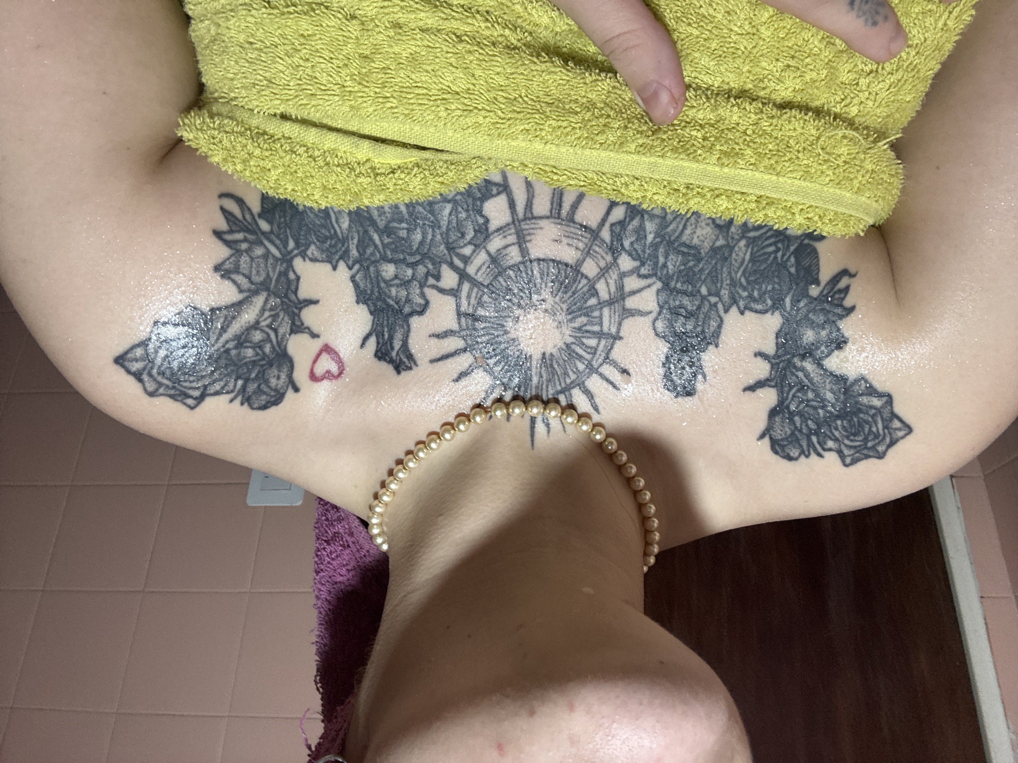 A close-up, high-angle shot shows a person from the chest up, their upper torso and shoulders covered in numerous black ink tattoos featuring roses and a compass or wheel-like design. The person is wearing a strand of pearls around their neck. A yellow towel is draped over the upper chest, near the shoulders, with a portion of a finger visible on the towel. The skin appears wet, possibly from a recent shower. In the background, there is a light purple towel and dark wooden panels or a door on the right side. The immediate surroundings suggest a bathroom setting, potentially in an indoor location. The information is insufficient to determine the location within Buenos Aires, Argentina.