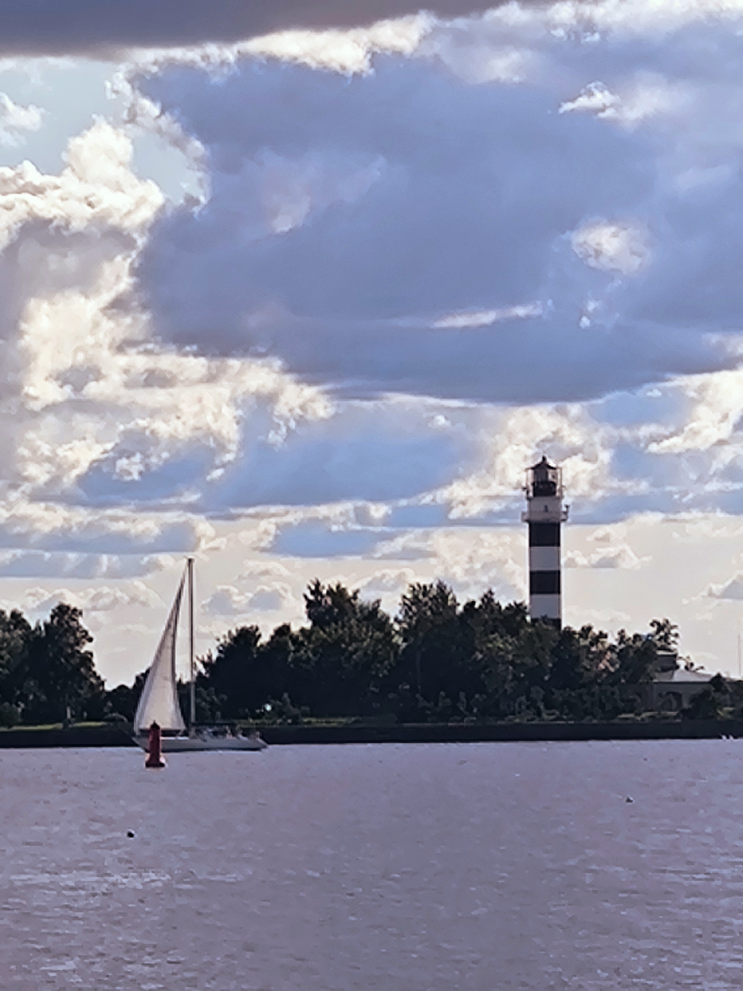 A sailboat with a white sail navigates a body of water, with a red buoy in the foreground. In the background, a lighthouse with black and white horizontal stripes stands amidst a cluster of trees. The sky above is filled with dramatic clouds, featuring a mix of dark gray and bright white. The scene is set against the backdrop of a lush green shoreline. This appears to be the Daugava River in Riga, Latvia, given the distinctive lighthouse and its common placement on the river.