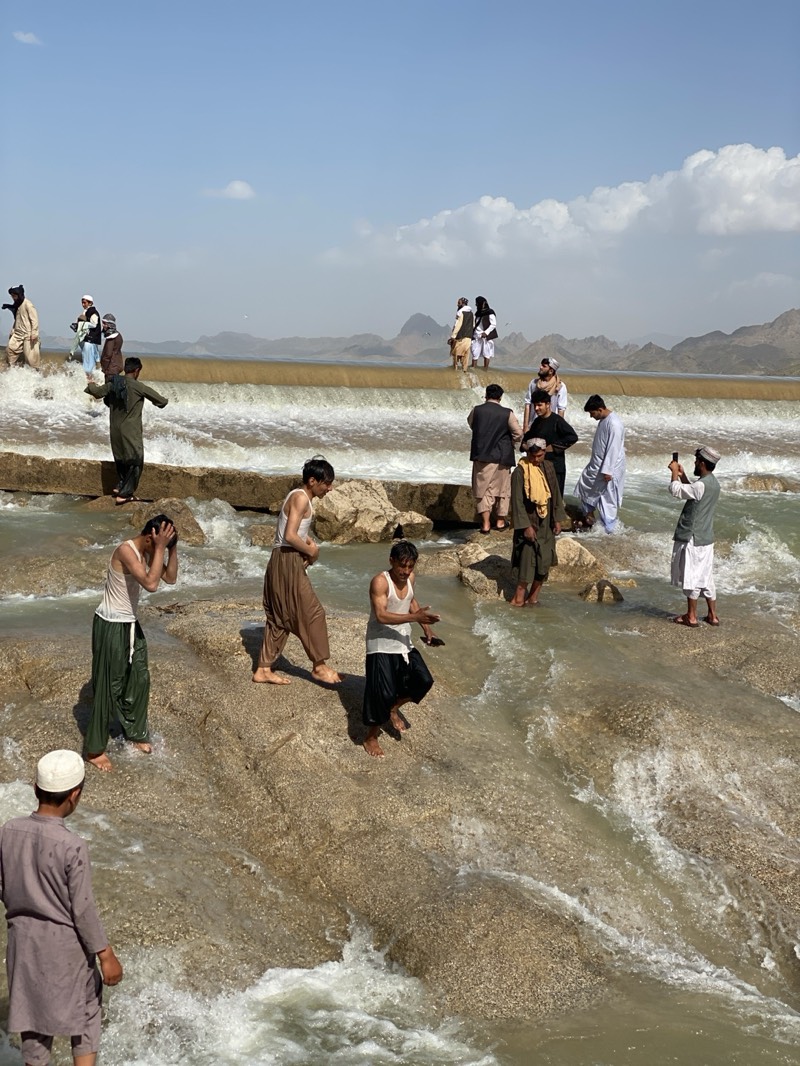 The image depicts a group of men and boys gathered at a rocky, shallow river or waterfall. Most of the individuals are wading in the water or standing on the rocks. Some are wearing traditional Afghan clothing, including turbans, loose pants, and long shirts. One man is holding a phone, appearing to take a photograph. In the background, a low dam or barrier creates a waterfall effect. Mountains are visible in the distance under a partly cloudy sky. The location is Kim, Afghanistan.