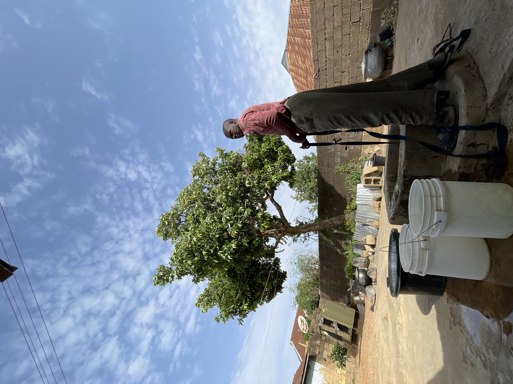 The image, taken in Jos, Nigeria, captures an outdoor scene dominated by a man engaged in drawing water. The man, appearing middle-aged with a bald head, wears a reddish-brown long-sleeved shirt and dark green or grey trousers. He leans forward, gripping a long, dark handle, suggesting he is operating a hand pump or a similar mechanism to retrieve water.
To the man's right, in the foreground, several plastic buckets are arranged on the ground: two white ones and one black bucket partially obscured beneath a white one. The ground appears to be concrete or packed earth, with wet patches indicating recent water spills.
A large, lush green tree with dense foliage stands prominently to the left of the man, under a bright blue sky filled with scattered, wispy white clouds. In the background, various buildings made of stone or brick are visible, featuring typical corrugated metal roofs. One building has a small, circular satellite dish attached, and a small, unreadable sign. Power lines or wires stretch across the upper left quadrant of the frame.
The strong sunlight and clear sky indicate it is daytime, likely late morning or early afternoon. The overall scene suggests a routine activity in a residential or communal area. No specific emotions are discernible on the man's face, which is focused on his task.