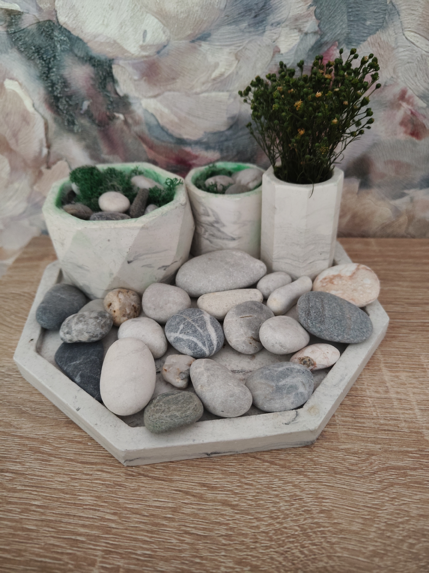 In Придорожный, Russia, on a wooden surface, a hexagonal concrete tray holds a collection of smooth, light-colored stones. Three small concrete planters are arranged behind the tray. Two of these planters are filled with green moss and smaller stones, while the third contains a dried, bushy plant with small yellow flowers. The background features a textured wall with a painterly, abstract design in shades of white, grey, blue, and pink.