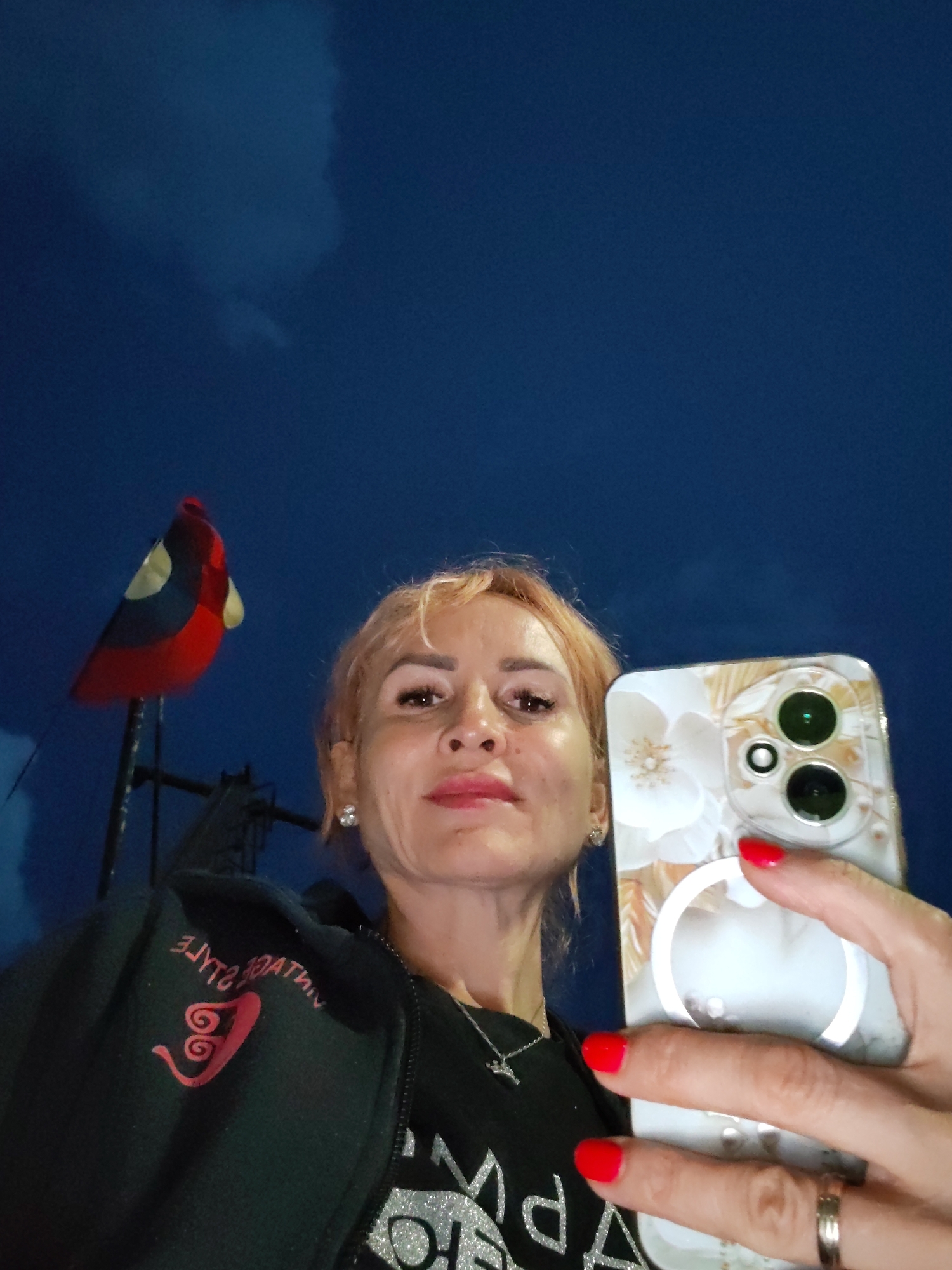 A selfie taken at dusk against a dark blue sky with faint cloud formations. A woman with blonde hair, wearing a black zip-up jacket over a sparkly shirt, holds a white floral-patterned phone in her right hand. Her fingernails are painted red. A silver ring is visible on her left hand. To the left of the woman is a flag on a pole, appearing to be red, black, and yellow. The image does not contain any discernible landmarks or features that would indicate a specific location within the city of Rybinsk, Russia.