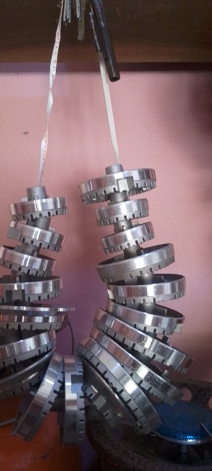 The image depicts two stacks of metal ring-like objects, possibly clutch plates or gears, suspended by strings. The objects are shiny and metallic, with notched edges. They are stacked on top of each other, forming cylindrical shapes. One string appears to be a measuring tape, with visible markings. The background is a pinkish wall. The scene is likely an indoor workshop or storage area. The presence of these specific mechanical parts suggests a location involved in automotive repair or manufacturing, which could be present in Jalingo, Nigeria, a city known for its commercial activities and informal workshops. The lighting is somewhat dim, suggesting artificial light or a shaded area.