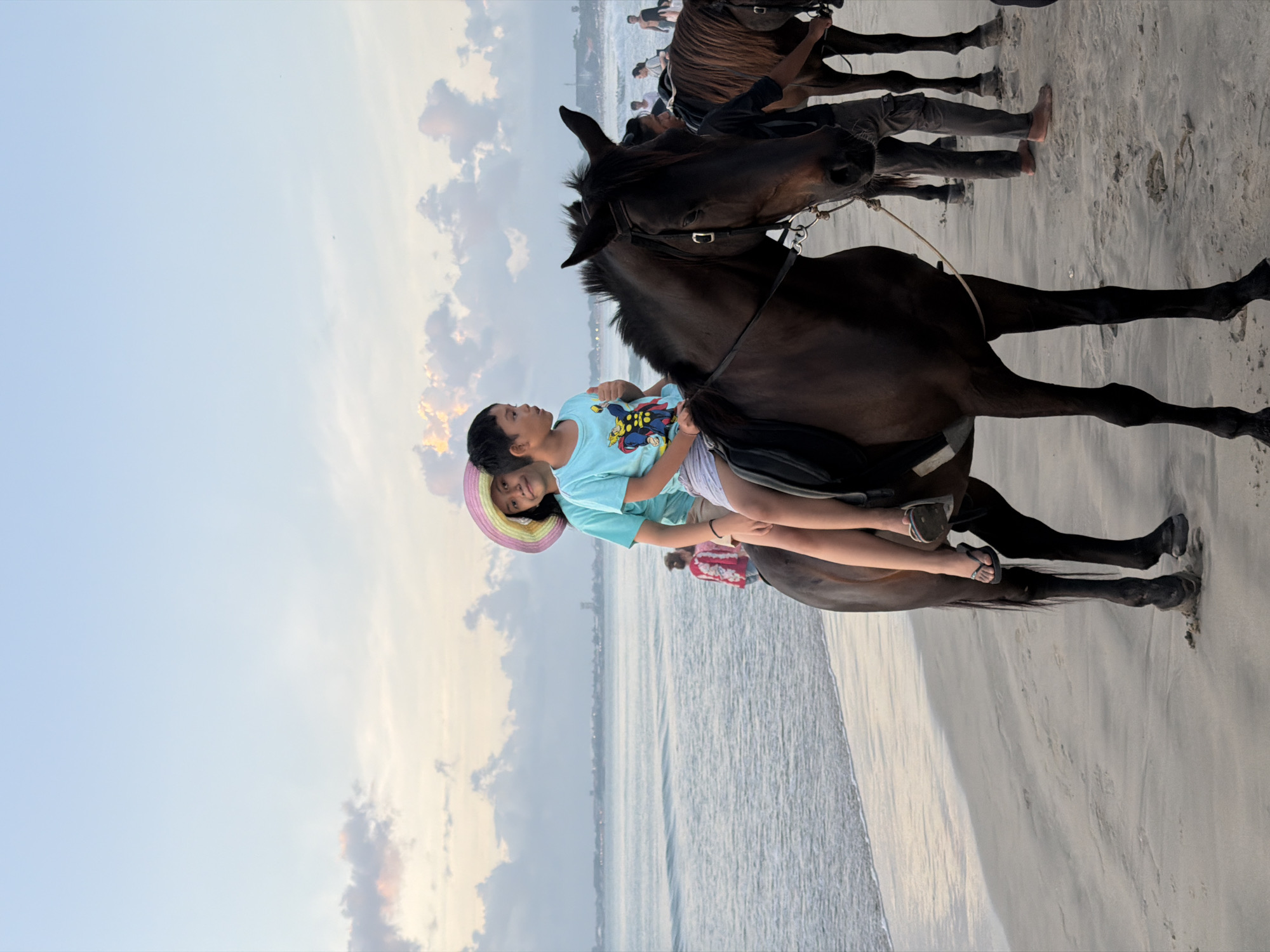 A medium-level shot shows a light-skinned child in a multi-colored wide-brimmed hat and light-blue t-shirt with cartoon figures holding onto a second light-skinned child seated on a dark-brown horse. The horse is facing the viewer’s right, and the back of the horse is nearest to the viewer. The sea and sky are in the background. Other people are seated on horses behind the focal horse. The children are on the beach in Kuta Selatan, Indonesia.