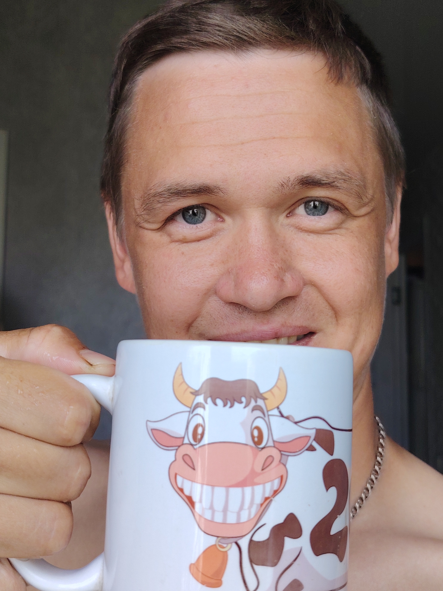 A close-up shot features a man with fair skin and short brown hair, holding a white mug with a cartoon cow design. He is looking towards the camera with a subtle smile and blue eyes. The mug has a large, cartoonish cow face with big teeth and horns, and the cow's body is visible on the side of the mug. The man is wearing a silver chain necklace. The background is blurred and appears to be an indoor setting, possibly a home. The lighting suggests it is daytime. The image was taken in Chaikovsky, Russia.