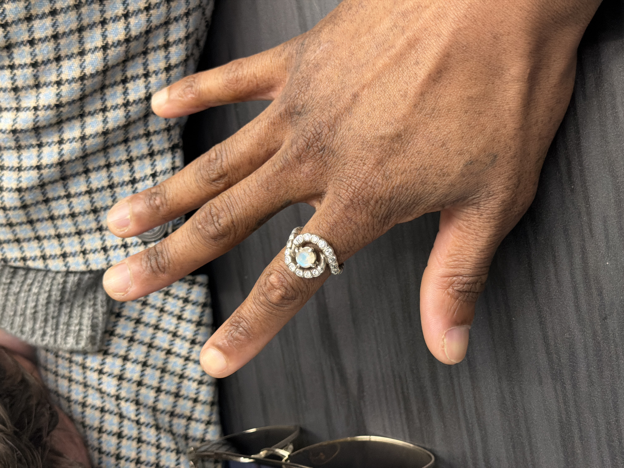 A close-up shot of a Black person's hand adorned with a silver ring on the ring finger. The ring features a central round stone surrounded by smaller stones in a spiral design. The person's hand is positioned against a background of a gray surface and a blue and gray patterned fabric. The lower portion of a pair of glasses is visible in the foreground. The image lacks any information to identify the specific location in the city of London, United Kingdom.