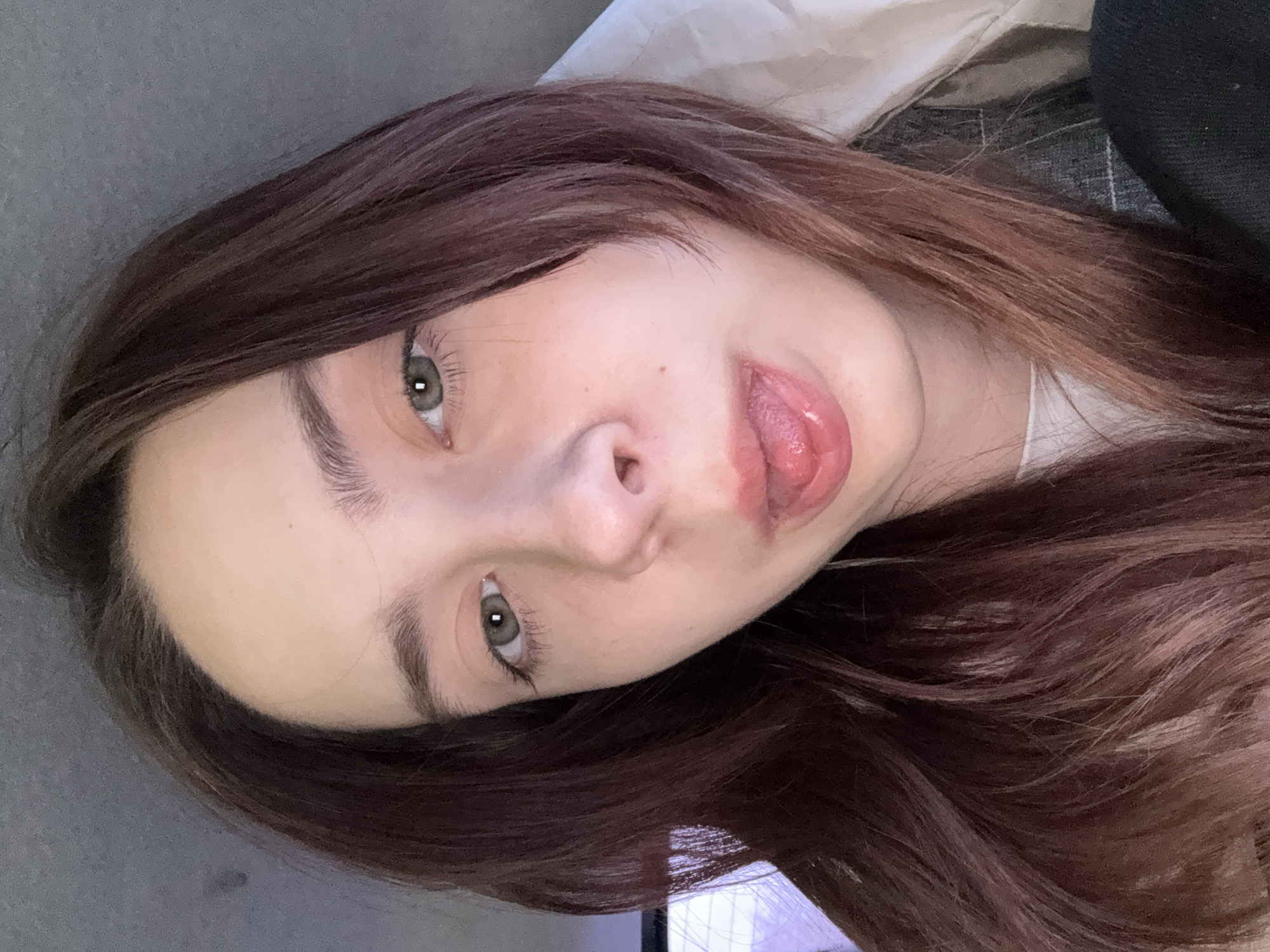 A close-up, rotated view of a young woman's face. She has fair skin, brown hair, and light green eyes. Her mouth is slightly open, with her tongue visible. The background is out of focus, showing a blend of gray and white tones, suggesting indoor lighting. The location and city of Chaikovsky, Russia, are not determinable from the image.