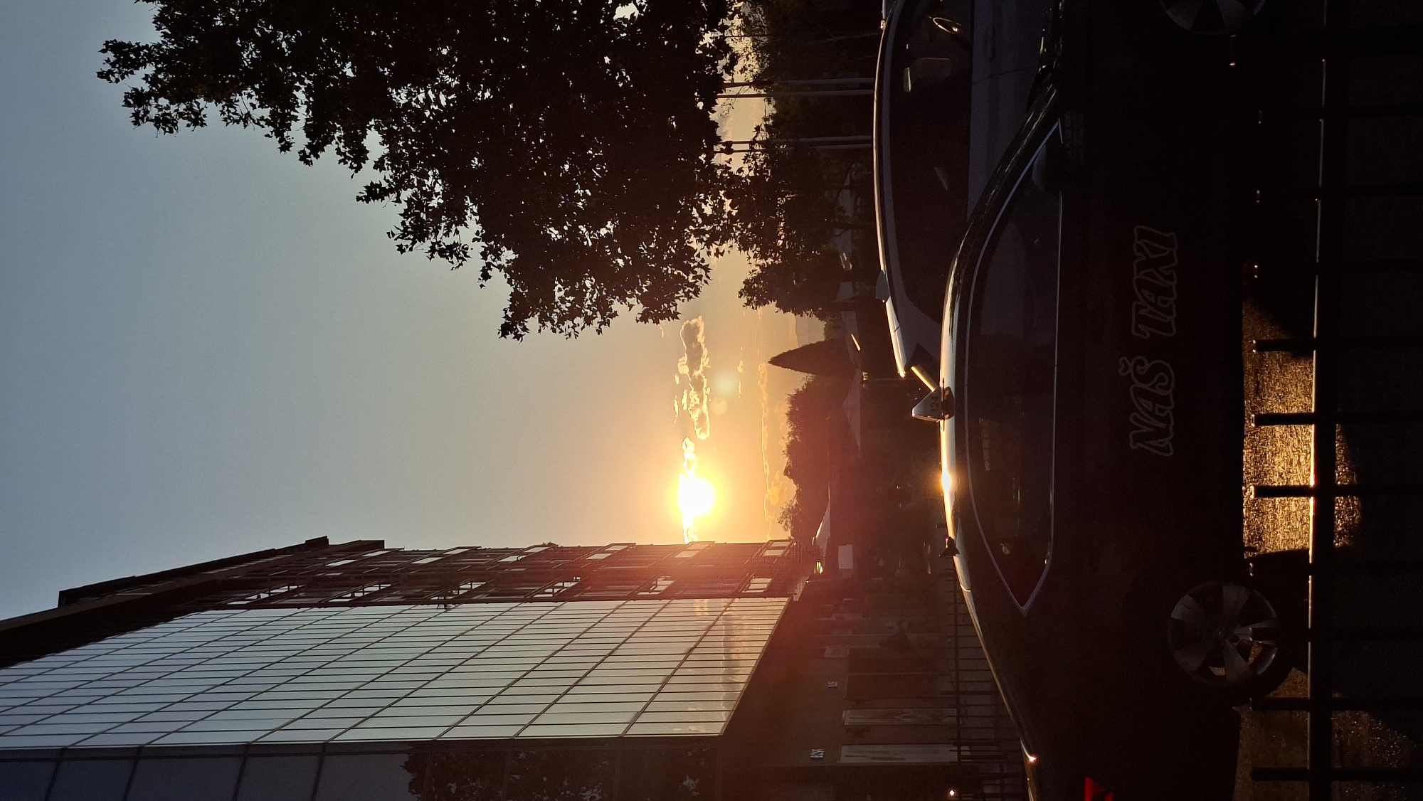 The image depicts a street scene at sunset in Podgorica, Montenegro. The sun, a bright orange orb, is low in the sky, casting warm light and long shadows. To the left, a modern, multi-story building with a glass facade reflects the sunlight. In the foreground, the silhouettes of two cars are visible, the one closer to the camera being a dark-colored taxi with the text "NAŠ TAXI" on its side. The taxi has a light on its roof and its wheels are visible. The ground appears wet, with reflections of the setting sun. Trees and distant hills form the background, silhouetted against the vibrant sky. The overall atmosphere is tranquil, capturing the end of the day in an urban setting.