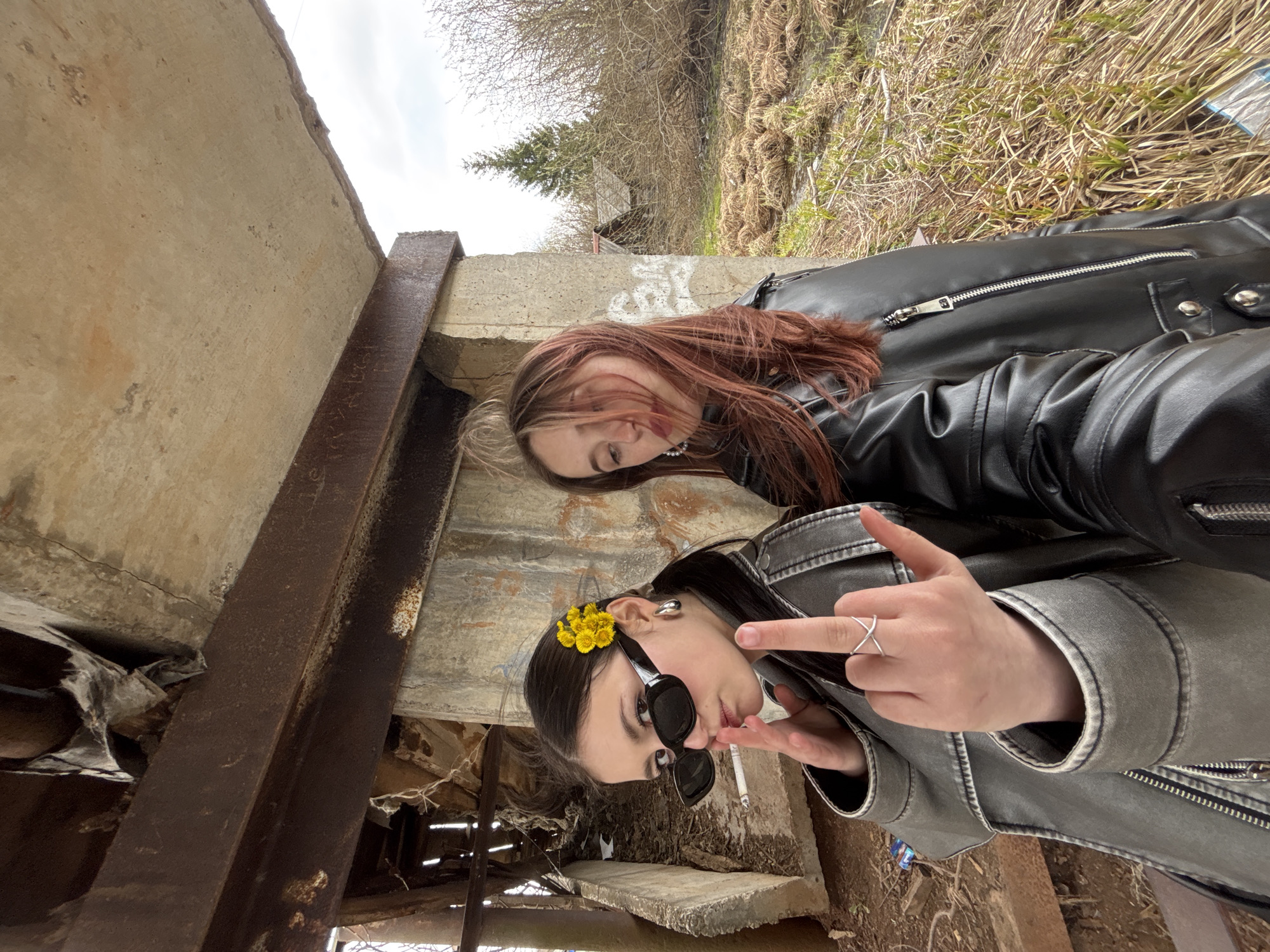 The image displays two young women posing in an outdoor setting characterized by industrial decay. The perspective is tilted, showing the subjects and surroundings at an angle.
In the foreground, a woman with dark hair is visible. She wears sunglasses, a dark gray or black leather jacket, and has several small yellow flowers tucked into her hair above her right ear. She sports a silver ear cuff and a silver 'X' ring on her left hand, which is raised in a middle-finger gesture towards the viewer. A cigarette hangs from her mouth.
Behind her, partially obscured, is another woman with reddish-brown hair, wearing a black leather jacket and a pearl necklace. She appears to be looking downwards, her expression less prominent than the foreground subject.
The setting consists of rough concrete walls and massive, rusted metal beams or girders, suggesting an abandoned or dilapidated structure like a bridge underpass or an old industrial site. The ground is covered with dry grass and sparse vegetation, with bare trees and shrubs visible against an overcast sky, indicating late winter or early spring. There are faint, illegible markings or graffiti on the concrete surfaces and some text-like etchings on the rusty metal beam, none of which are decipherable. The scene conveys a raw, somewhat rebellious aesthetic, fitting for a rural or semi-urban environment in Фоки, Russia, during daytime under cloudy skies.