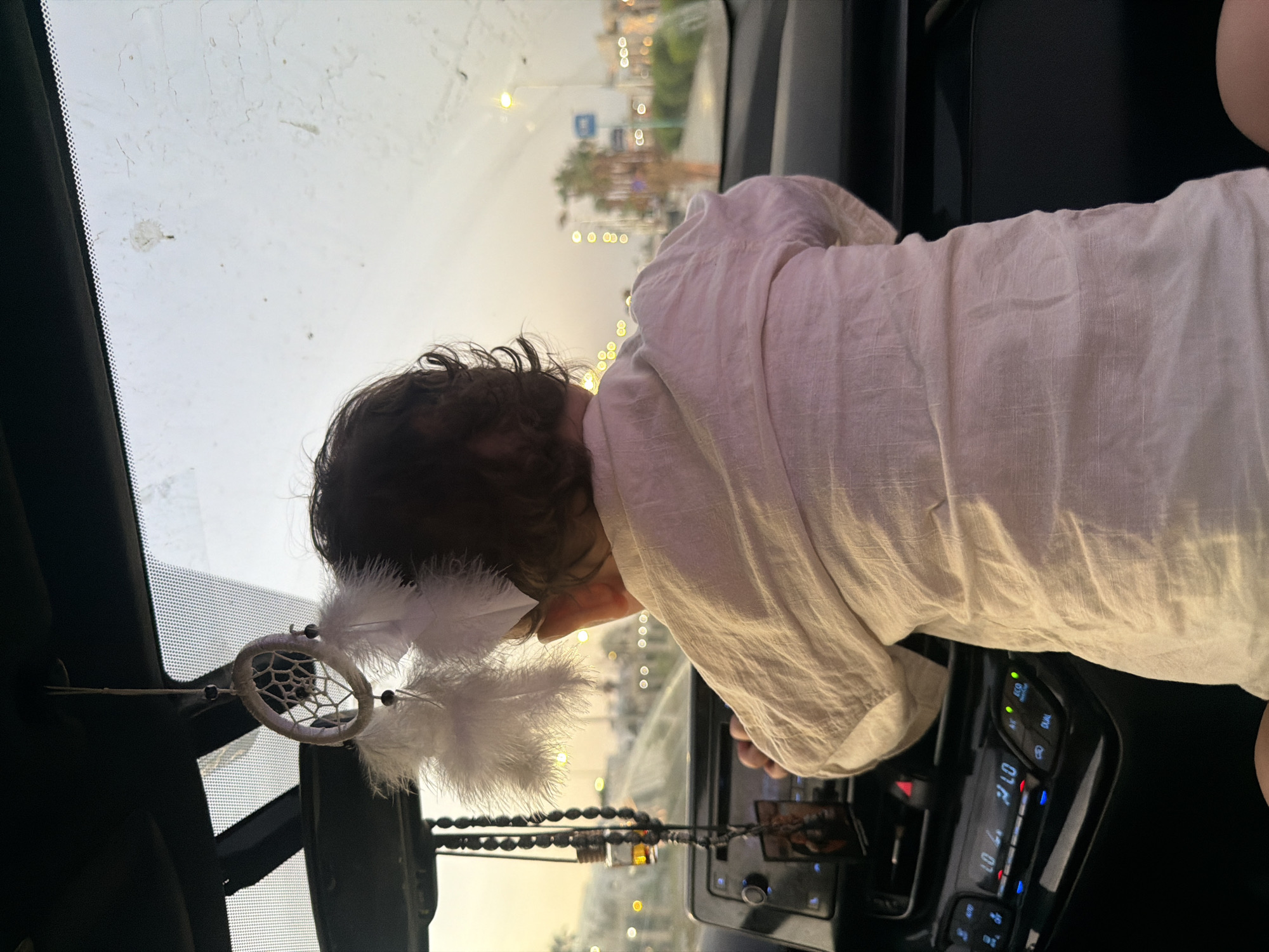 A rear view of a baby in a car in Sharm El Sheikh, Egypt. The baby, facing away from the camera, is wearing a white linen shirt and is sitting in the driver's seat, appearing to hold the steering wheel. The car's dashboard is visible, with its infotainment system displaying time and temperature. A dreamcatcher with white feathers hangs from the rearview mirror. Outside the car, streetlights and palm trees line a road, indicating an urban or suburban setting during dusk or dawn. The sky is hazy with soft, diffused light. The car's windshield shows some moisture or smudges. The scene conveys a sense of everyday life in Sharm El Sheikh.