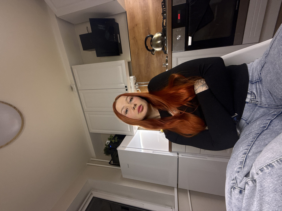 The image captures a young woman with long, flowing vibrant red hair, seated in what appears to be a home kitchen or dining area. She is looking directly at the camera with a neutral or slightly relaxed expression, displaying a nose ring (septum piercing) and prominent eyebrows. She is wearing a long-sleeved black top and light-wash blue jeans, with tattoos visible on her left forearm.
The background shows a modern kitchen with white upper and lower cabinets and a light wooden countertop. Above the stovetop, there is a black range hood. On the countertop, a dark-colored stovetop is visible, with a metallic electric kettle resting on it. Below the stovetop, an oven is integrated into the cabinetry, featuring a digital display that shows "00:40". To the left of the woman, a white refrigerator is partially visible, and next to it, on a lower shelf or cabinet, there's a potted plant with dark foliage.
The walls of the room are a light, possibly cream or beige, color. A circular light fixture with a wooden frame is partially visible on the ceiling in the upper left of the frame. In the lower left corner, a window reveals a dark exterior, suggesting it is nighttime. White pipes run horizontally along the wall beneath the window. The overall setting appears to be a casual indoor environment.