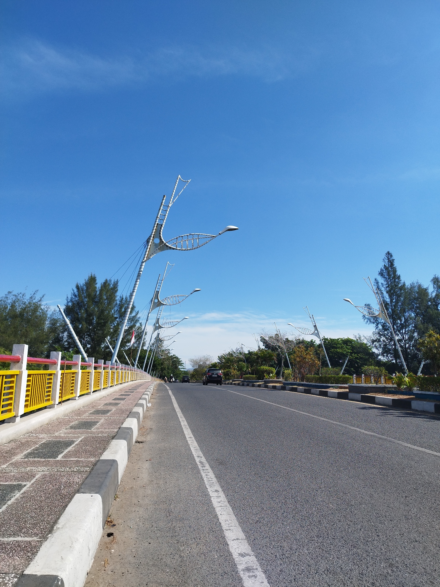 The image shows a street scene in Pango Raya, Indonesia. On the left side of the road, there is a sidewalk with a yellow and red railing and a sidewalk made of textured concrete tiles. Along the sidewalk, a series of distinctive streetlights with decorative, wing-like structures are visible, extending upwards and curving. Behind the railing and sidewalk, trees and greenery are present. The road itself is asphalt with white lane markings and is mostly empty, with a single black car visible in the distance. The sky is clear and bright blue with a few wispy clouds. The overall impression is a well-maintained urban street with unique architectural elements, likely a coastal or park area given the clear sky and roadside greenery.