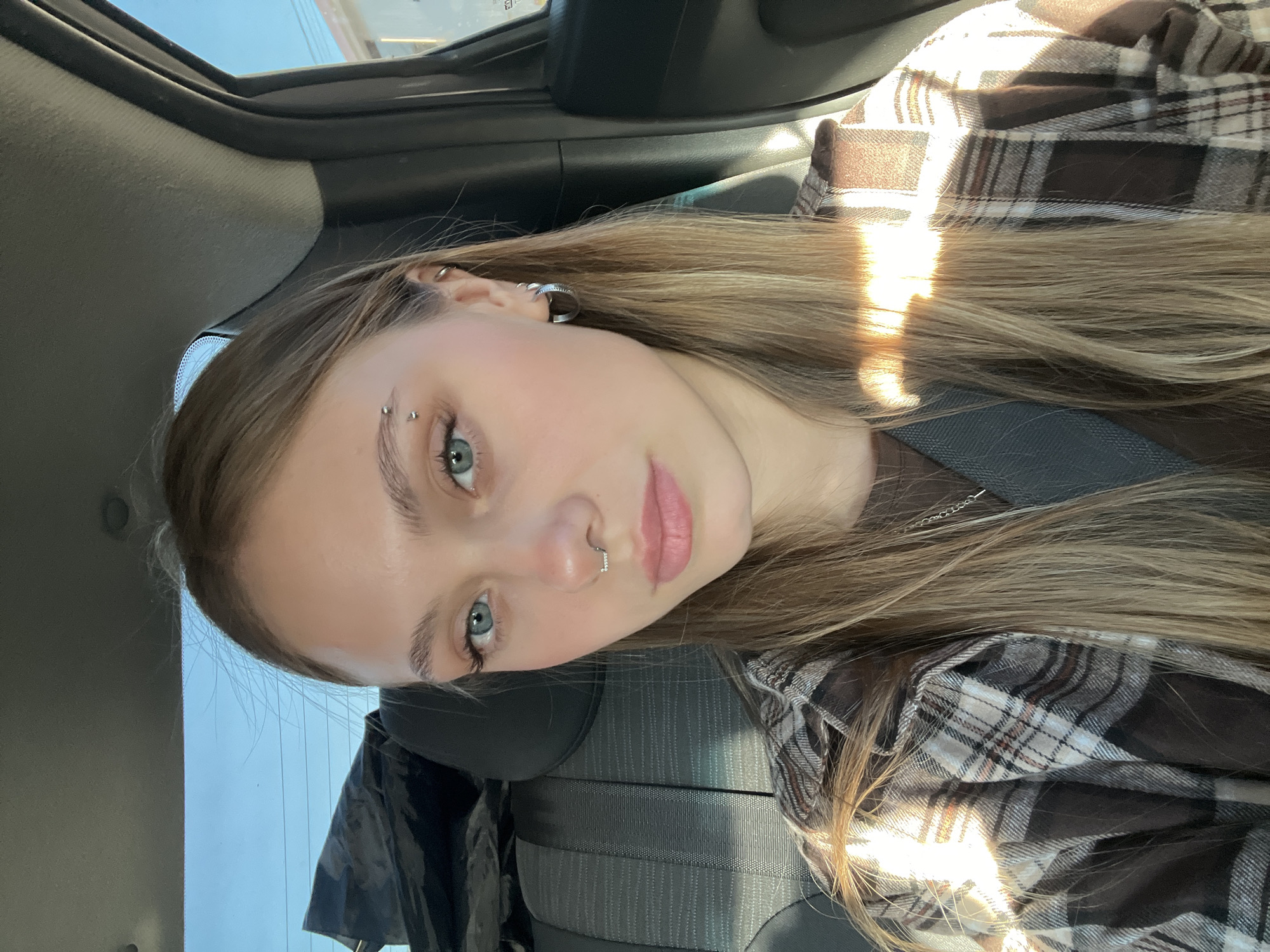A young woman with fair skin, long blonde hair with highlights, and blue eyes is positioned in the passenger seat of a car. She is wearing a brown and tan plaid shirt and a brown shirt underneath. She has a silver hoop earring in her upper ear, an eyebrow piercing, and a nose piercing. A seatbelt crosses her chest. The interior of the car is visible, including the seat and the car's ceiling. The background shows a glimpse of the outside, suggesting a bright, possibly sunny day. The location and specific details related to the city of Chaikovsky, Russia are unknown in this image.
