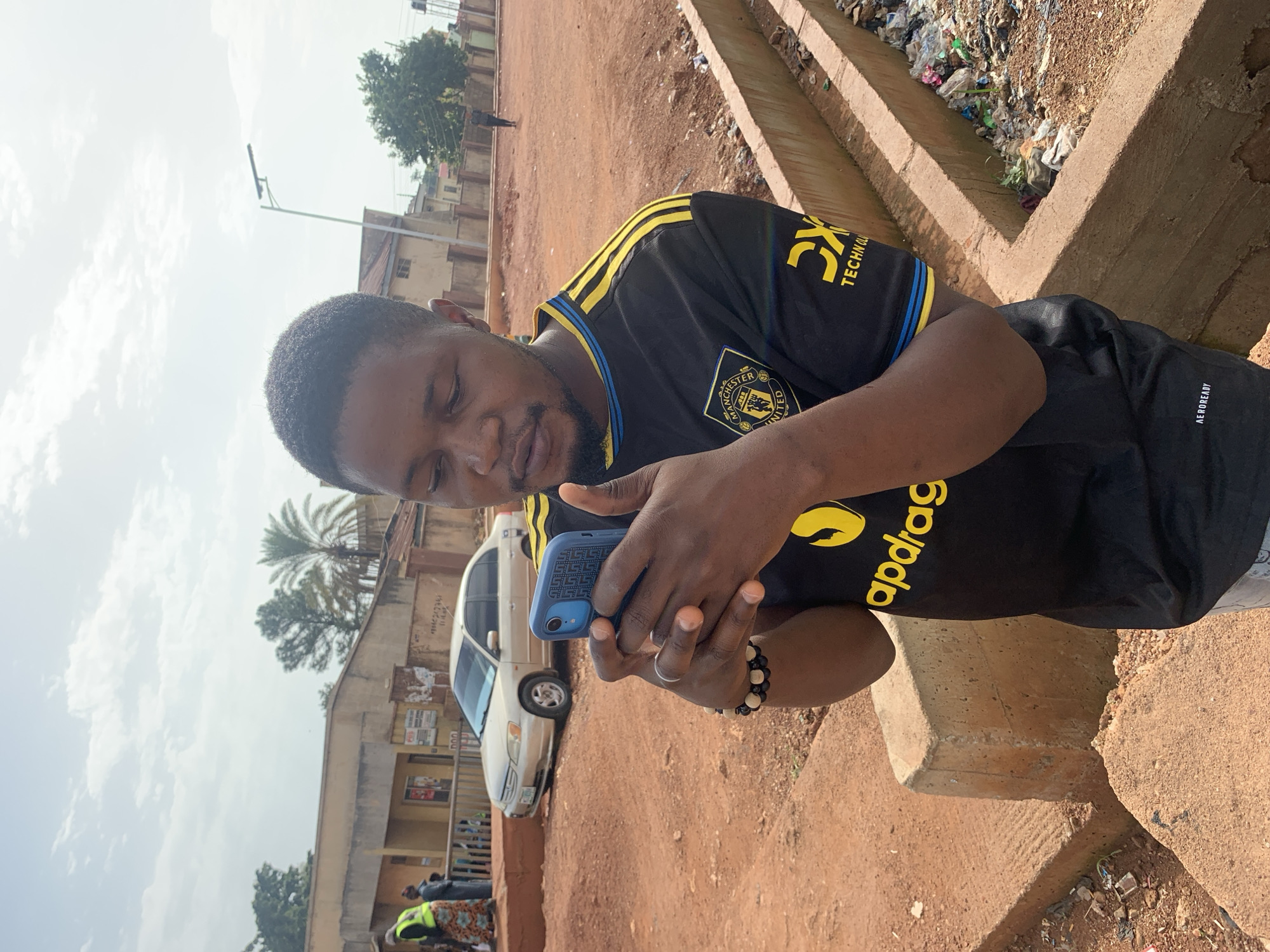 A young Black man in a black Manchester United jersey with yellow trim is holding a blue smartphone in his hands and looking at it. He is standing in an outdoor environment that appears to be a public space. The man is wearing a beaded bracelet on his left wrist. In the background, a light-colored sedan is parked. There are buildings and a street lamp visible. The ground is a reddish-brown dirt. The image was taken in Gura Top, Nigeria.