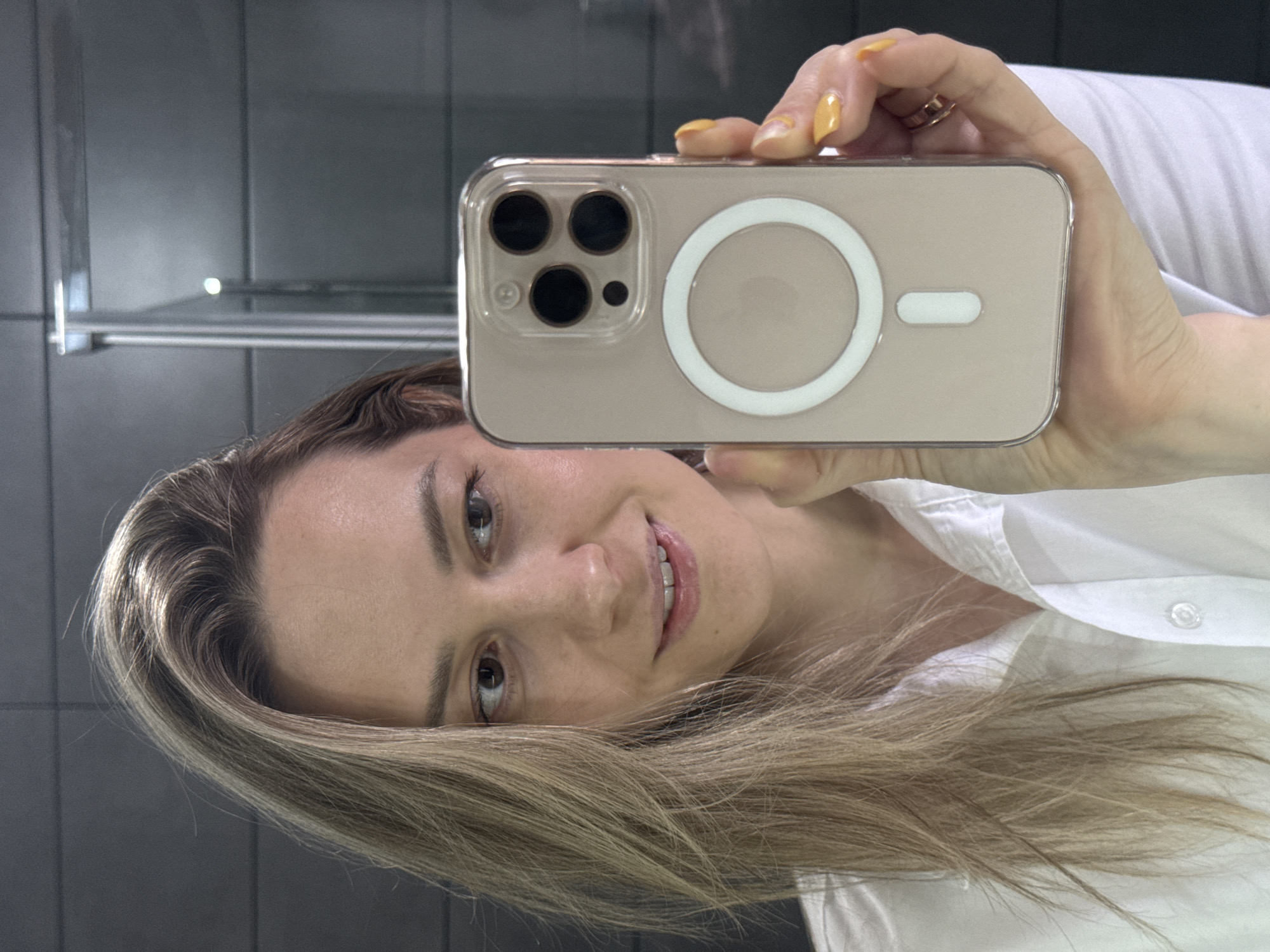 A woman with blonde hair is taking a selfie with a cell phone. The phone is in a clear case with a MagSafe charging ring on the back. She is wearing a white button-down shirt. The background is a dark gray tiled wall with a metal shelf. The location is inferred to be in the city of Chaikovsky, Russia.