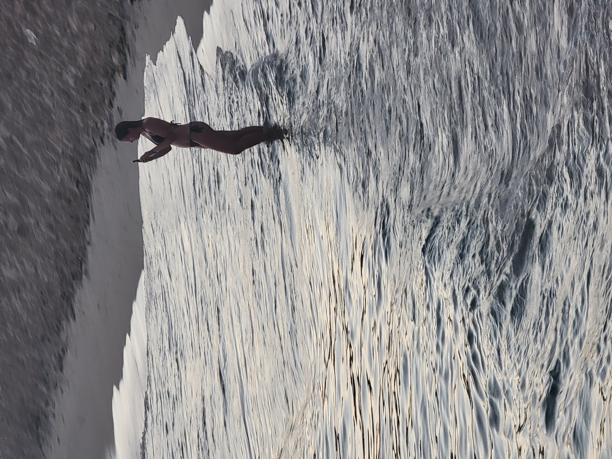 A woman stands in the shallow water at the edge of the ocean on a beach, holding a smartphone and taking a picture. She is wearing a black bikini. The water has small waves. The sand and the edge of the water are visible in the background.
