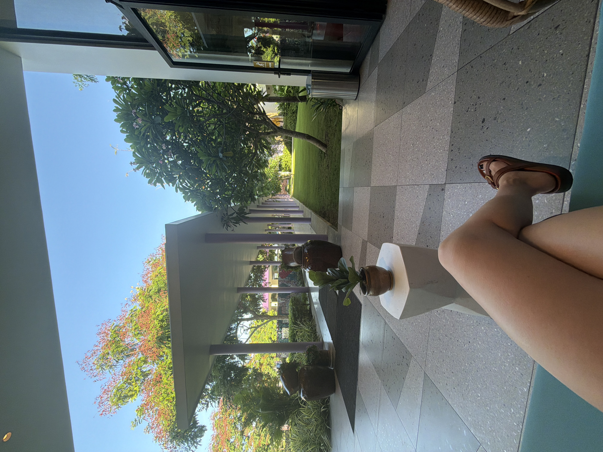 The image depicts an outdoor covered walkway with a tiled floor, leading to a grassy courtyard. The walkway is lined with purple columns and features large decorative ceramic pots. In the foreground, a person's legs are visible, crossed and resting on a teal surface. The person is wearing brown sandals. A small potted plant sits on a white geometric stool near the person. Beyond the walkway, a lush green lawn is visible, with trees and some buildings in the distance. The sky is clear and blue. The scene appears to be in a tropical or subtropical location. While the image is described as being taken in Dong Ninh Hoa Ward, Vietnam, there are no specific visual cues within the image to confirm this location.