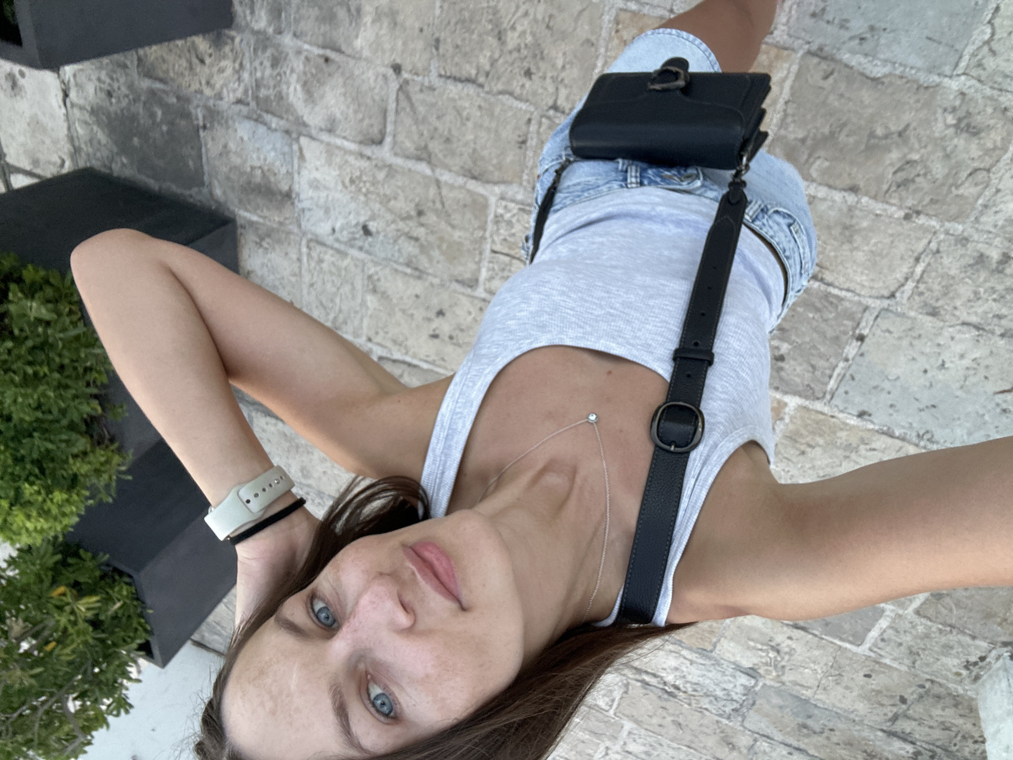 The image features a young woman with blue eyes and brown hair, lying down outdoors. She is wearing a light grey tank top, denim shorts, and a black crossbody bag. A light-colored smartwatch is visible on her left wrist, along with a thin silver necklace with a small pendant. The background consists of a stone-tiled walkway and some greenery. The lighting suggests it is daytime, likely sunny. The woman's expression appears relaxed or thoughtful. The scene is set in Budva, Montenegro.