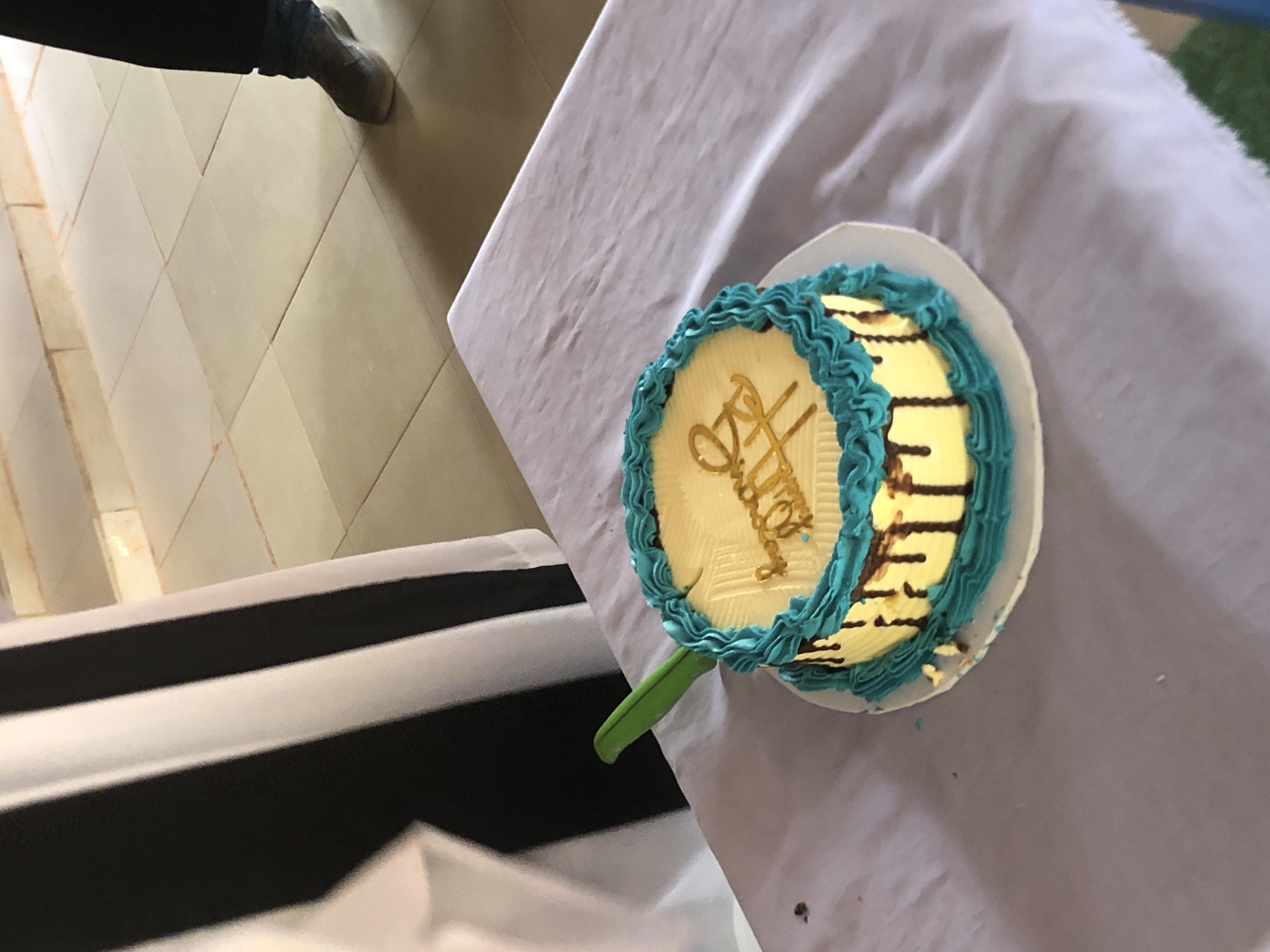 The image shows a partially eaten round cake with light yellow frosting, decorated with gold lettering and a turquoise-colored frosting around the edge. The cake is placed on a white plate atop a light purple tablecloth, which is on a table. A green plastic knife is inserted into the cake. To the left of the table is a black and white striped object. A person's leg and shoe are visible in the top left corner, suggesting the photo was taken from above. The location cannot be determined with the given information.