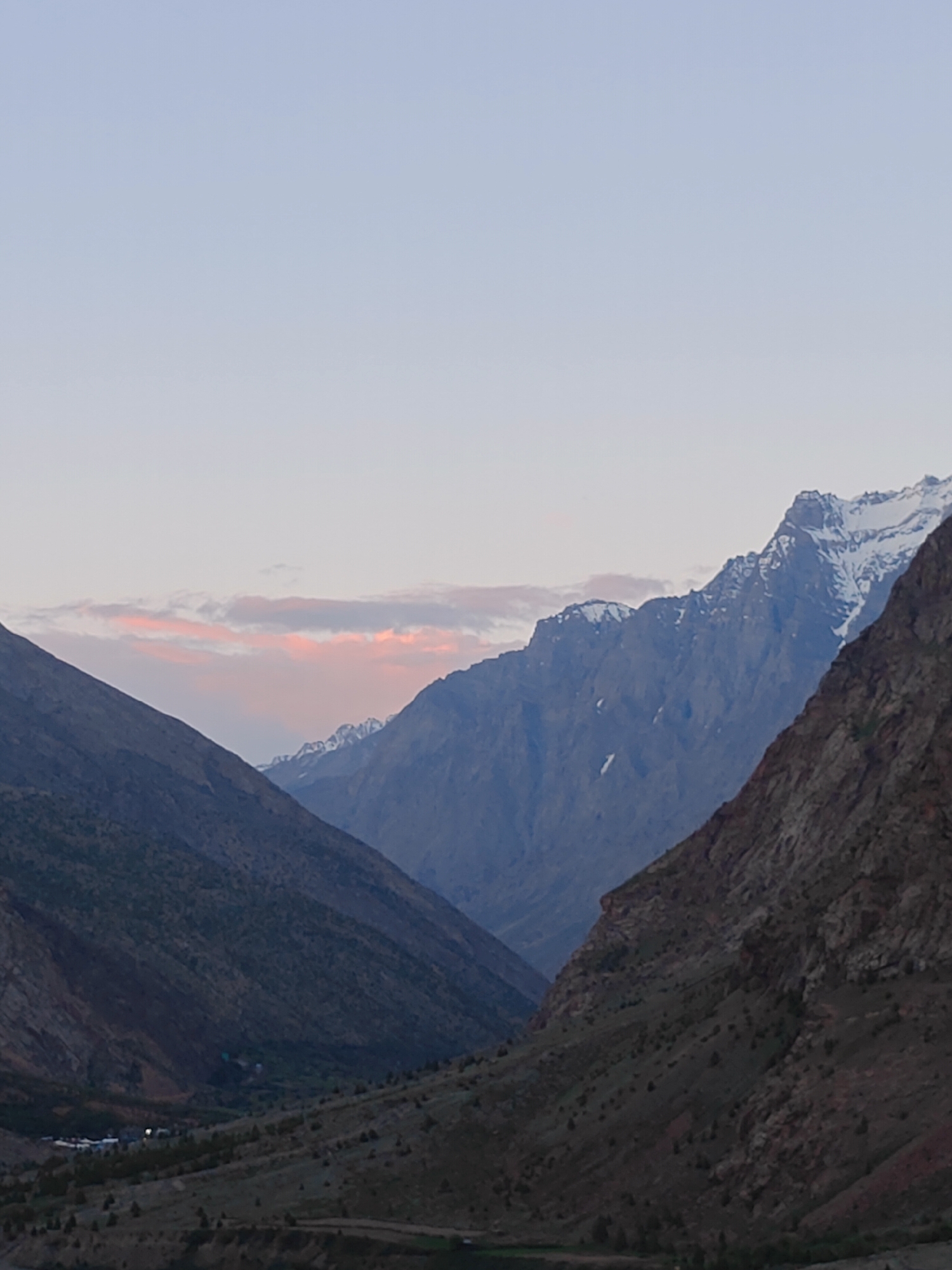 The image depicts a mountainous landscape, possibly during twilight or dawn, given the soft lighting and pastel colors in the sky. In the foreground, two large mountain slopes converge, creating a valley that recedes into the distance. The left slope is darker and appears to be covered in sparse vegetation. The right slope is a reddish-brown, rocky mountainside with scattered green shrubs.
Further in the distance, beyond the initial slopes, are more rugged mountains. Some of the higher peaks are capped with snow, reflecting the dim light. A soft band of pink and orange clouds stretches across the sky horizontally, just above the distant mountains.
In the valley floor, visible in the lower part of the image, there appears to be a small settlement. Several buildings are clustered together, and some lights are visible, suggesting human habitation. The area around the settlement is dotted with trees and appears to be more cultivated than the surrounding slopes. The overall impression is one of a remote, natural environment with a small human presence nestled within. The location is identified as Gemur, India.