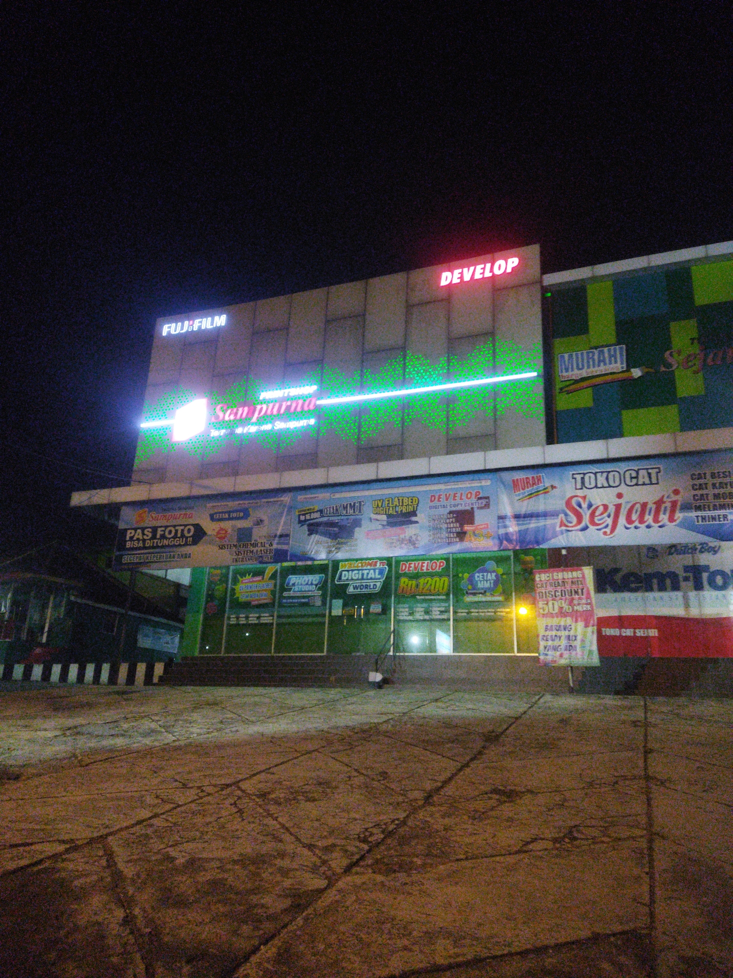 The image shows the exterior of a commercial building at night, likely a retail establishment, located in Wonogiri, Indonesia. The building features several illuminated signs advertising various services and products. Prominent among these are signs for "FUJIFILM" and "DEVELOP," indicating a photographic and digital printing service. Another large banner advertises "TOKO CAT Sejati," a paint store, with additional text about discounts on "CUCI GUDANG" (warehouse cleaning) and "BARANG READY MIX YANG ADA" (ready-mixed goods available). The building's facade is modern, with some sections exhibiting a grid-like pattern. Green and blue colored panels are visible on the upper right portion of the building. The ground in front of the building is paved and appears to be a walkway or parking area, illuminated by the building's lights and casting shadows. The overall scene suggests a busy commercial street or complex operating at night.