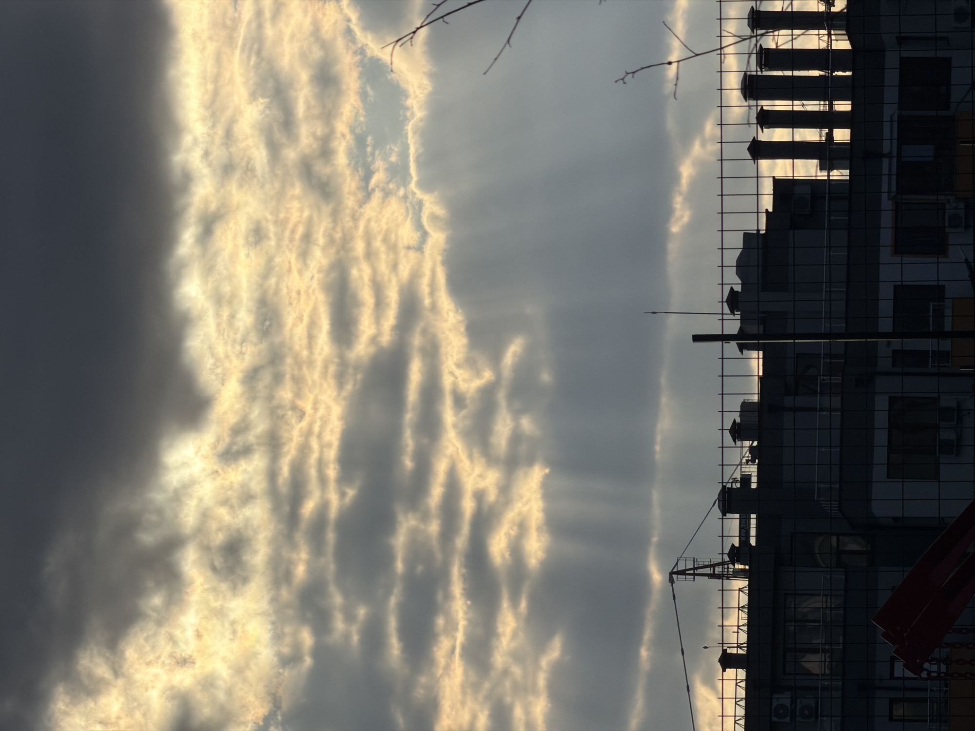 The image displays a dramatic urban sunset or sunrise over Varna, Bulgaria, characterized by striking atmospheric lighting and silhouetted city elements. The sky, vibrant and dynamic, occupies the upper two-thirds of the frame. On the left, bright golden sunlight pierces through textured clouds, casting distinct crepuscular rays that fan out across the sky. These sun-kissed clouds form intricate, wavy patterns, while the clouds towards the right appear darker and denser, creating a stark contrast.
Along the lower third of the image, the dark silhouettes of multi-story buildings define the cityscape. These structures feature numerous windows, some reflecting faint light, and prominent rooftop elements like chimneys, vents, and air conditioning units. A grid-like fence or scaffolding partially covers the buildings, particularly towards the right side. On the far left, the distinct outline of a construction crane is visible. Bare tree branches extend into the upper sky from the left and right edges. Faint, partially legible text, possibly "WWW.MU..." or similar, can be seen reflected in one of the building's windows. The overall scene evokes a sense of serene beauty, with the dramatic natural light highlighting the urban landscape at dawn or dusk.