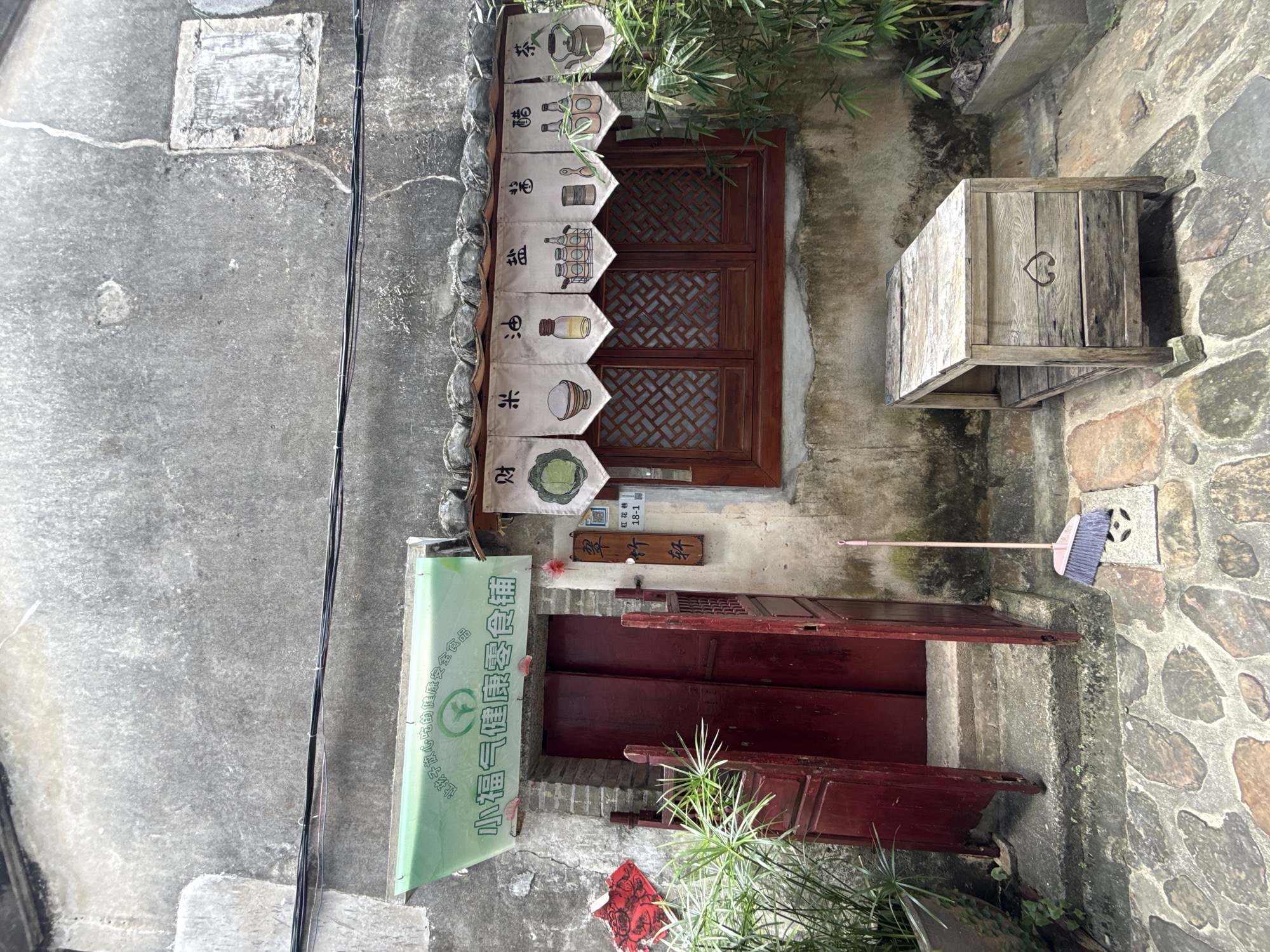 A full shot captures the exterior of a small shop with a weathered concrete facade in Longgang District, China. The entrance features a pair of dark red wooden doors, one ajar, with a wooden gate. Above the doorway hangs a green banner with Chinese characters and a circular green logo. To the right of the entrance is a wooden-framed window with intricate geometric patterns. A valance made of small fabric banners, each depicting a different Chinese character and illustration related to food ingredients like cabbage, rice, oil, salt, sauce, vinegar, and tea, hangs above the window. A wooden bench with a carved heart detail sits in front of the window. In the foreground, stone paving leads to the entrance, and a pink broom rests near a drainage grate. Green plants are visible on the left and right sides of the scene. The overall impression is of a traditional, possibly rustic, establishment.