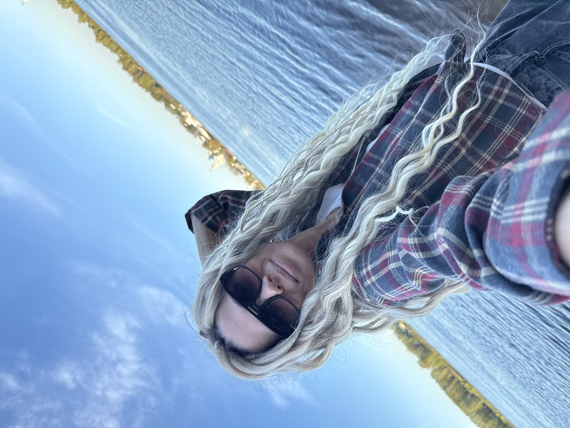 A selfie taken by a person with blond hair and sunglasses, wearing a plaid shirt and dark jeans. The background shows a body of water, likely a river or lake, and a clear blue sky with some clouds. In the distance, a cityscape with trees is visible. This image may be taken near a body of water near the city of Rusanovo, Russia.
