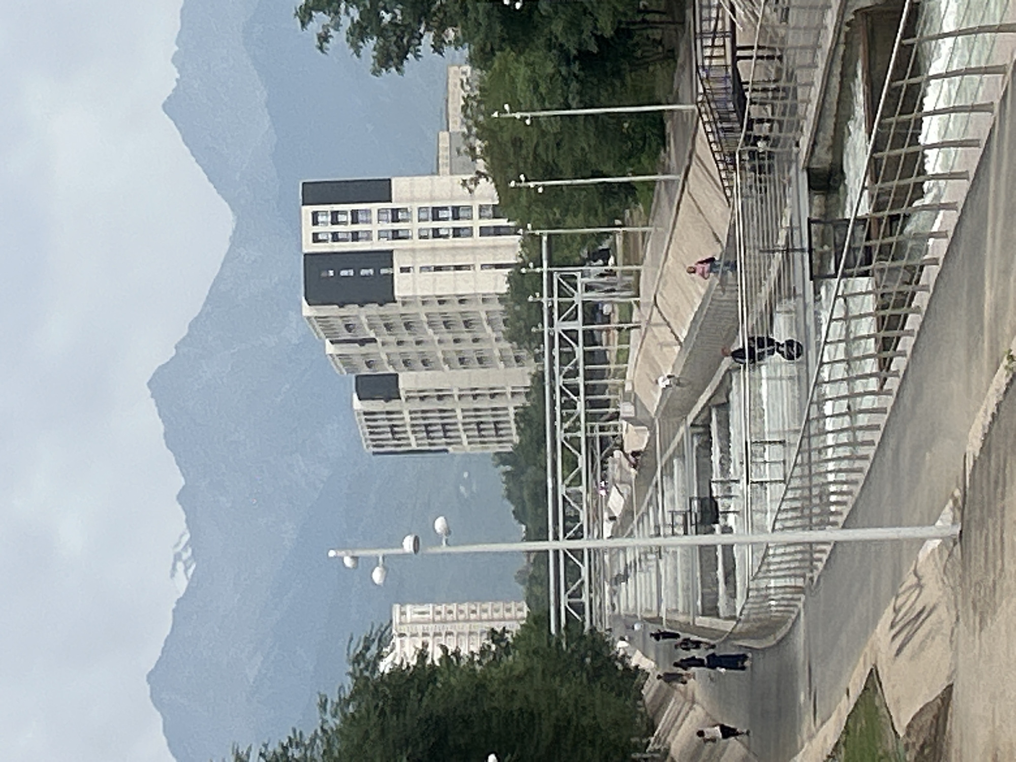 This is a photograph of the Almaty River rapids park in Almaty, Kazakhstan. The image captures a wide view of the park, with a focus on the man-made rapids and surrounding urban and natural elements. In the foreground, a paved walkway curves along the side of the rapids, bordered by a metal railing. Several people are visible on the walkway, some walking and others standing. The rapids themselves are a prominent feature, with water flowing through a series of concrete channels and structures. Bridges span across the rapids at various points, and more people can be seen on these bridges.
In the midground, modern apartment buildings rise, characterized by their white and dark grey facades and numerous windows. Trees and greenery are interspersed between the buildings and along the edges of the park, adding a natural element to the urban landscape.
In the background, a mountain range looms, its peaks appearing misty and somewhat indistinct against the overcast sky. The overall atmosphere is one of an active recreational space within a city that is nestled against a dramatic natural backdrop. The lighting suggests it is daytime, possibly overcast given the soft, diffused light and muted colors.