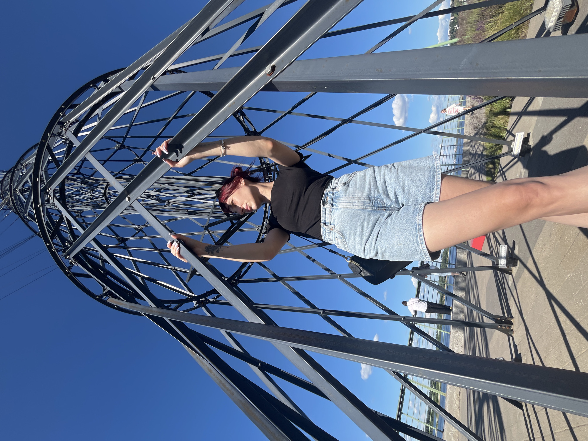 The media file shows a young woman posing on a metal structure, possibly a viewing platform or observation tower, under a bright blue sky with scattered clouds. She is wearing a black t-shirt and denim shorts, with her hair tied up. Her arms are outstretched, gripping the metal beams of the structure. The setting appears to be outdoors, with a body of water and a distant shoreline visible. The sun is shining brightly, casting shadows on the ground. There is another person visible in the background, further down the walkway. The overall impression is one of a leisurely day spent exploring a scenic location. The image is taken from a low angle, emphasizing the scale of the metal structure and the vastness of the sky. The location is identified as Nizhny Novgorod, Russia.