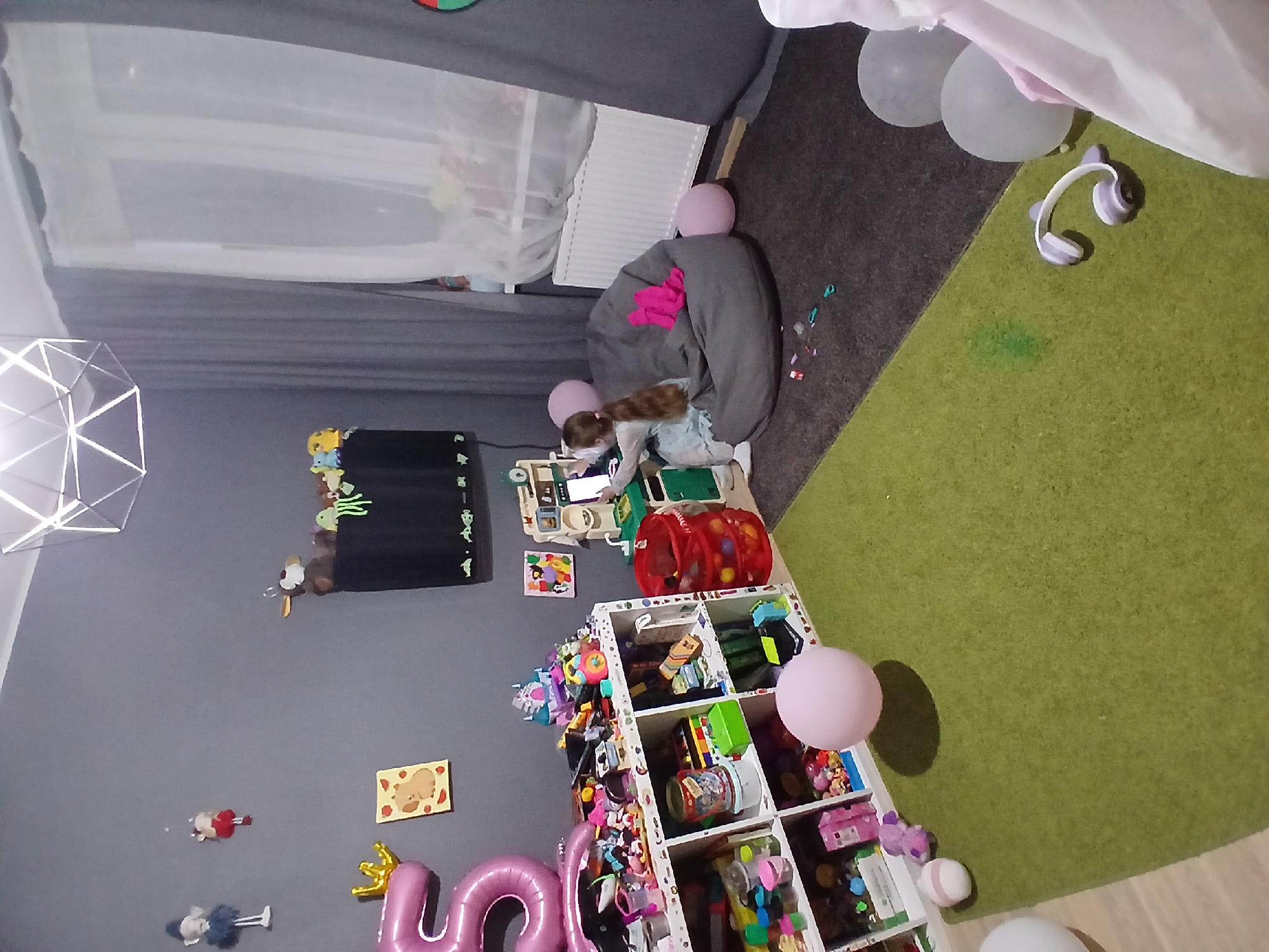 This is an indoor scene, likely a child's bedroom or playroom. The room is decorated in shades of gray and has a large green rug. On the left side of the room, there is a shelf unit filled with toys and art supplies. A large pink "5" balloon is also visible, along with a golden crown balloon. In the center of the room, a young girl with long brown hair is sitting on a large gray beanbag chair, playing with a toy kitchen set. She is wearing a light blue dress. There are several pink balloons scattered around the room. The room is illuminated by a modern geometric light fixture on the ceiling and natural light coming from a window covered by sheer white curtains. The overall impression is a playful and lived-in space. The image was taken in Yekaterinburg, Russia.