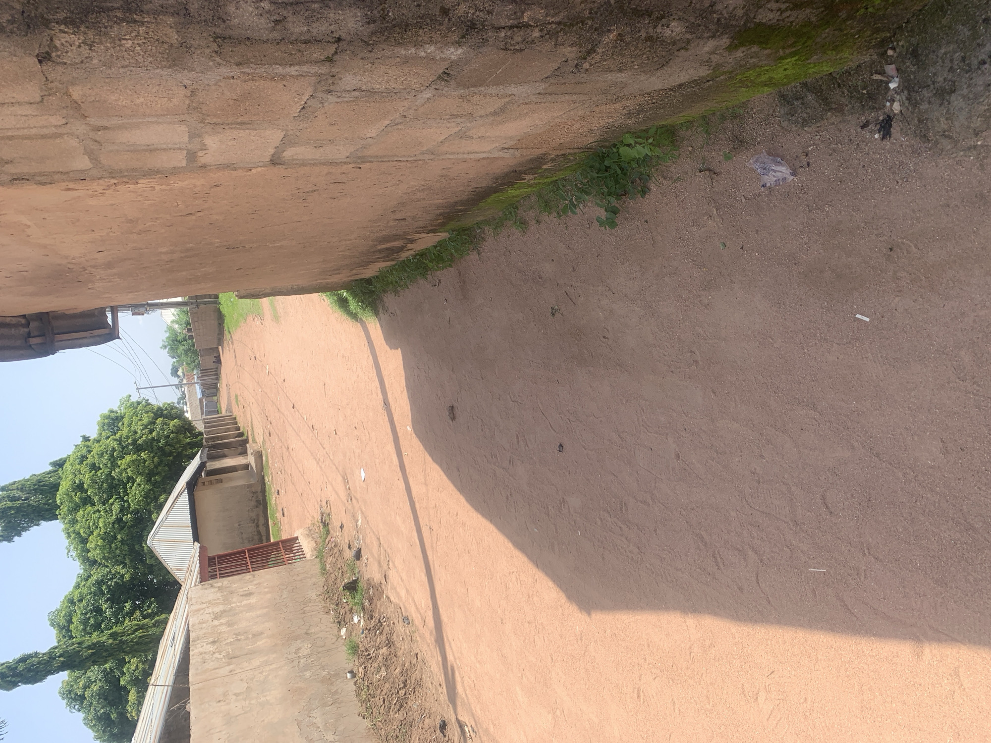 This is a medium-angle, outdoor, eye-level shot of a dirt road in Jalingo, Nigeria. The road is a reddish-brown color and appears to be made of clay or packed dirt. On the left side of the road, there are a series of low-lying buildings with corrugated metal roofs. Some of these buildings have beige or light brown walls, and one has a red metal gate. There are also some scattered piles of dirt and debris along the side of the road. Tall, slender trees with green foliage are visible in the background behind the buildings. On the right side of the road, a rough, textured wall made of concrete blocks is prominent. This wall has a reddish-brown, weathered appearance and some patches of green moss or algae growing at its base. The ground next to this wall is also covered in dirt with some small green plants and a piece of clear plastic litter. The lighting suggests it is daytime, with a clear sky and shadows cast across the road.
