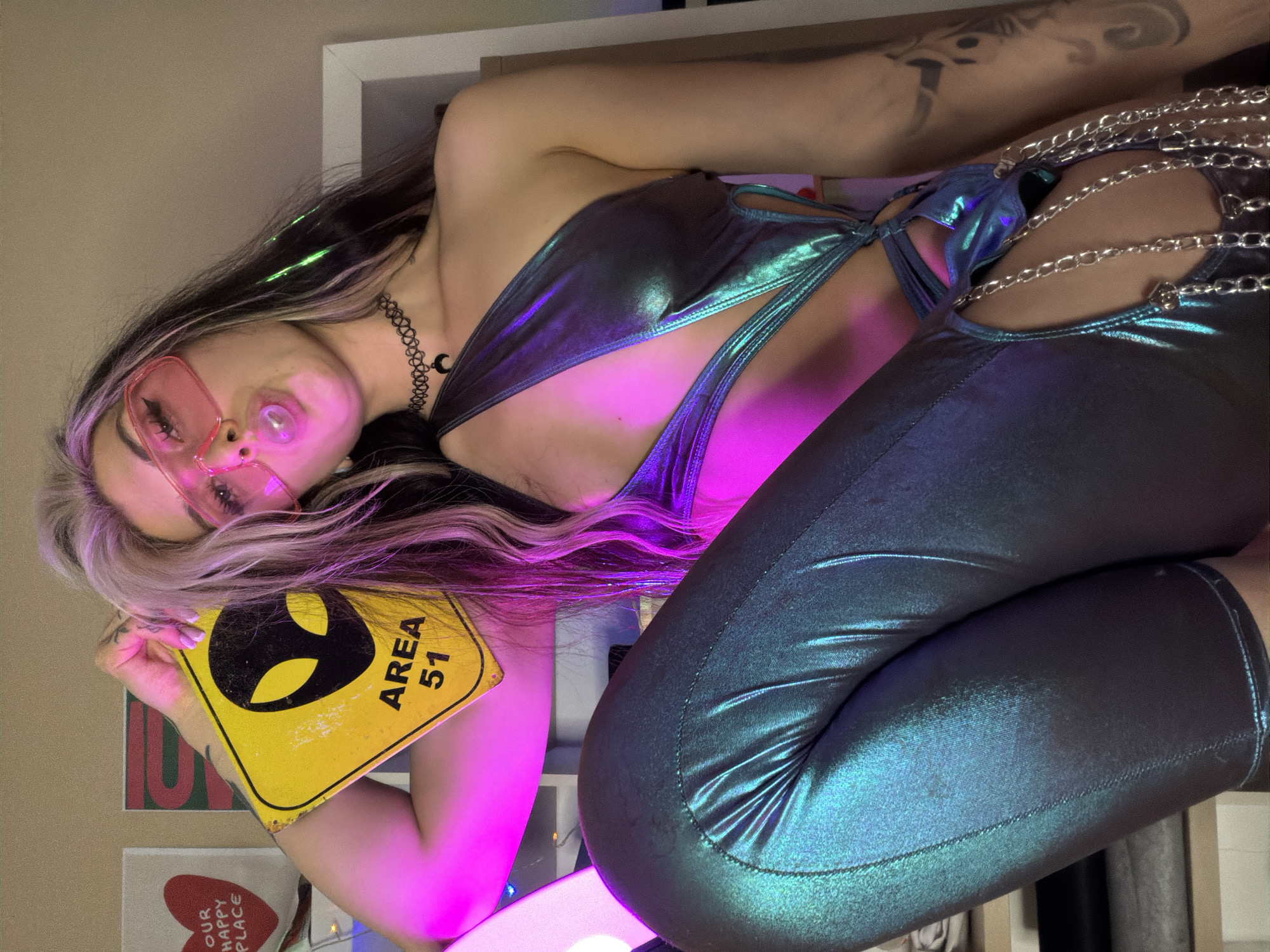An individual is depicted in an indoor setting, illuminated by prominent pink and green neon lighting. The subject is a female with long, dark hair featuring light-colored streaks, wearing large, rectangular, translucent pink sunglasses. She is blowing a bubble with pink chewing gum. A black choker necklace with a silver crescent moon charm is visible around her neck. She wears a metallic, iridescent, skin-tight bodysuit or jumpsuit in shades of teal, blue, and purple, featuring a significant cut-out on the left side of her torso and thigh, from which multiple silver chains dangle. A dark, abstract tattoo is visible on her right forearm. Her left hand, with light-colored fingernail polish, holds a yellow, diamond-shaped sign with a black alien head silhouette and the text "AREA 51". She is posed with her left leg bent and elevated in the foreground, facing the camera. The background consists of a plain, light-colored wall. To the left, two pieces of artwork are visible: a white canvas with a red heart and "OUR HAPPY PLACE" text, and another, less distinct, abstract canvas. A white shelf or furniture frame is partially visible on the right. The specific location within the city of Budva, Montenegro, is not discernible from the provided image.