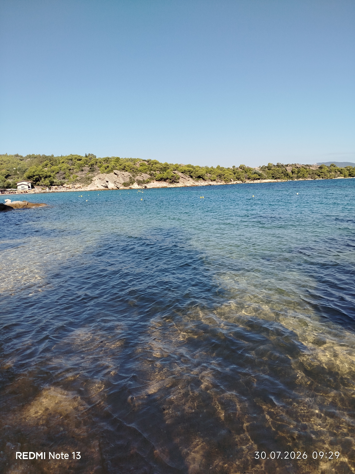 The image displays a clear, blue sky over a tranquil body of water. The foreground features shallow, clear water revealing the sandy seabed. In the middle ground, a shoreline with rocky outcrops and lush green vegetation can be seen. A few small buildings, possibly vacation homes or beach facilities, are situated along the coastline, along with beach umbrellas and chairs. Several yellow buoys are visible in the water, marking a swimming area or indicating a boundary. The overall scene depicts a serene beach or bay setting, likely in Sithonia, Greece, given the characteristic landscape. The timestamp "30.07.2026 09:29" and the text "REDMI Note 13" are overlaid on the image, indicating the date, time, and device used for capture.