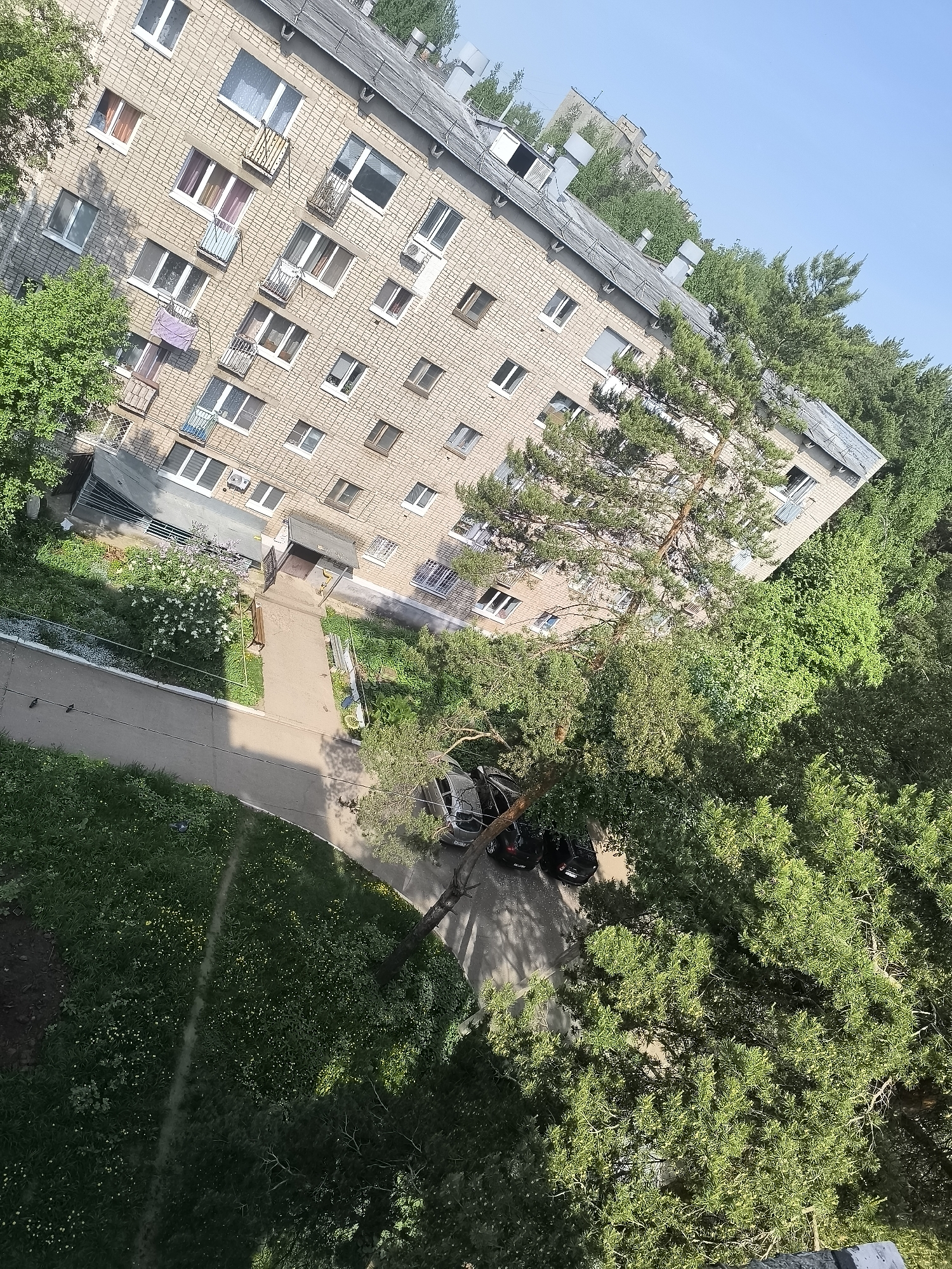 The image shows an aerial view of an apartment building in Unknown, Unknown. The building is made of light-colored bricks with multiple windows and some balconies. In front of the building, there is a paved walkway leading to the entrance, with grass and flowerbeds on either side. There are trees surrounding the building, partially obscuring it. A few cars are parked near the trees. The sky is visible in the upper right corner of the image.