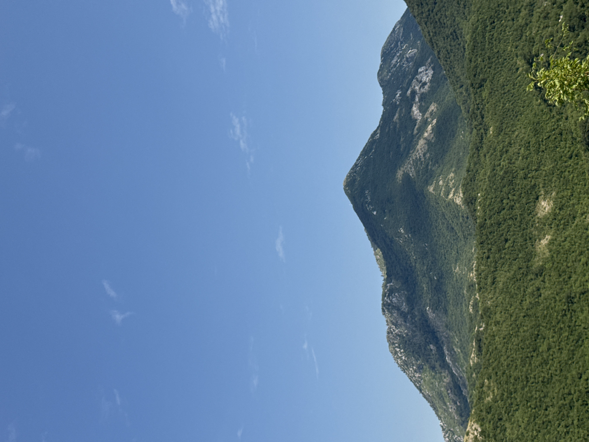 The image shows a panoramic view of a mountainous landscape under a clear blue sky with a few wispy clouds. The foreground is dominated by dense green forestation covering rolling hills. In the mid-ground, a larger, conical mountain rises, its slopes also covered in green vegetation, with some rocky, lighter-colored outcrops visible. The background consists of more distant, rugged mountains, also green and rocky. The scene appears to be a natural, undeveloped area, likely in a rural or wilderness setting. No people or man-made structures are visible. The location is identified as the city of Dragalj, Montenegro, a region known for its mountainous terrain and natural beauty.
