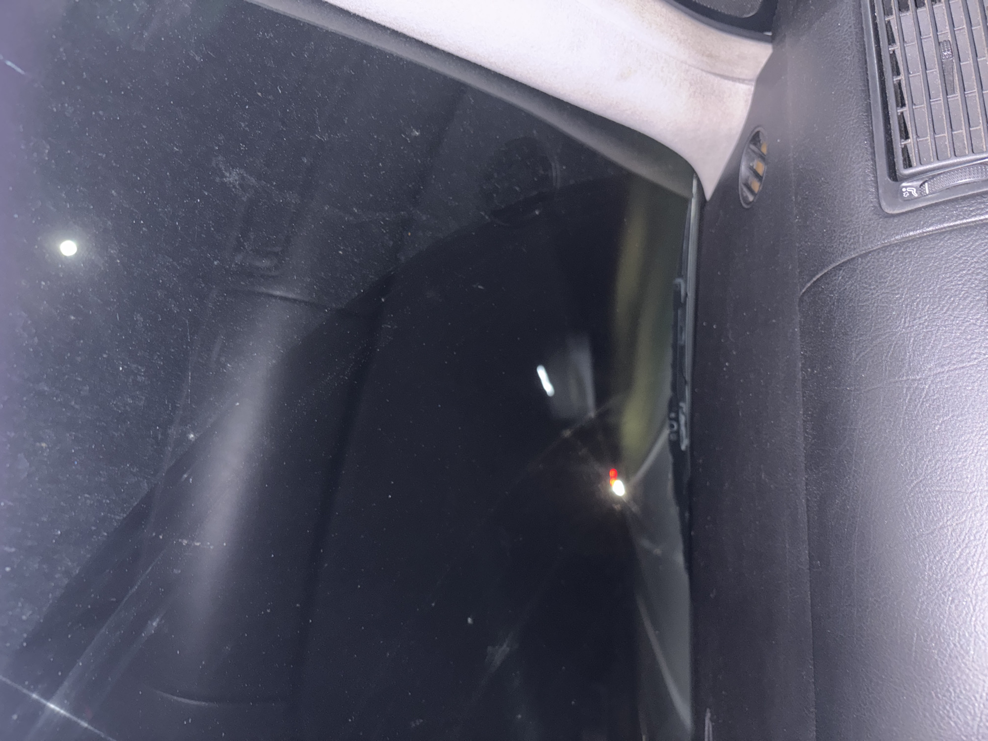 The image shows an interior view through a car window at night. The location is inferred to be Novo Leski, Bulgaria, based on the prompt context. The view is obscured by a dirty car window. A faint light source is visible through the darkness. A dashboard vent is visible on the right. The dashboard appears to be black plastic or leather. The headliner trim is visible in the upper section of the image. No people, actions, or interactions are visible. The environment is dark, with only reflected lights visible.