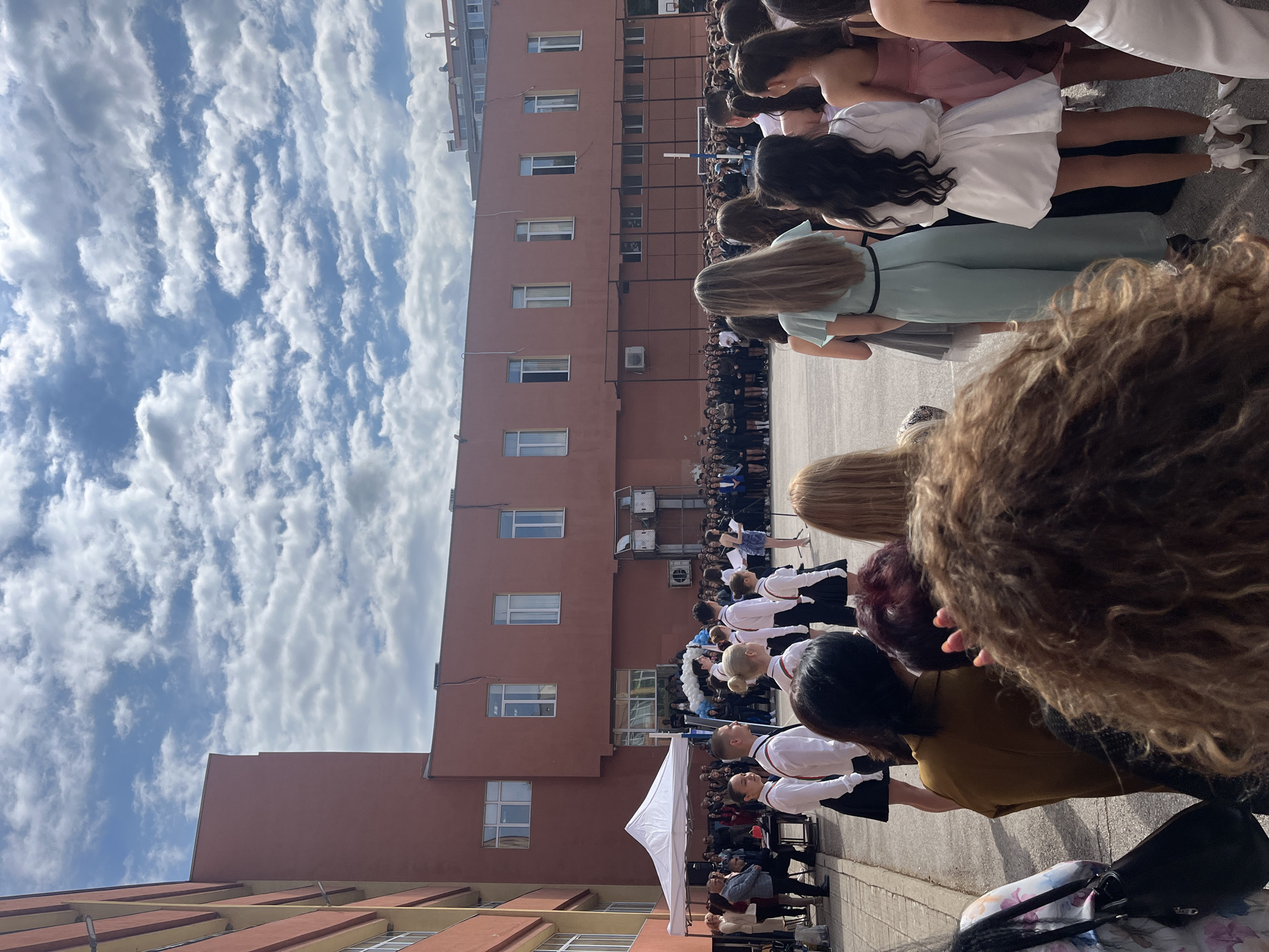 The image captures an outdoor event likely taking place in Blagoevgrad, Bulgaria. A large group of people, possibly students, are gathered in what appears to be a courtyard or open space adjacent to a tall, multi-story building, characterized by a reddish-brown facade and numerous windows. Some individuals are dressed formally, suggesting a special occasion. A white tent structure is visible, indicating an organized event. The sky is partially covered with clouds. The image is taken from within the crowd, focusing on the backs of people's heads, and capturing a range of hairstyles and clothing.