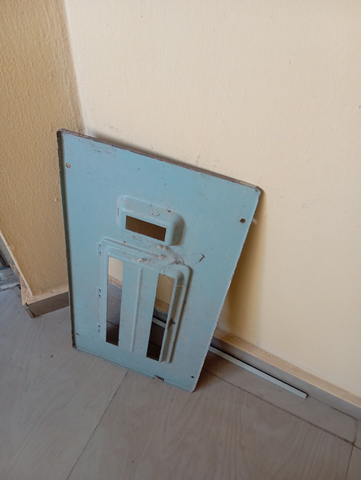 A light blue metal panel, likely a cover or a part of a larger appliance or structure, is propped against a wall in a corner. The panel has several rectangular cutouts of varying sizes. The wall behind it is painted in two colors: a lighter shade of yellow or cream on the right and a darker, textured tan or beige on the left, indicating a corner of a room. The floor appears to be tiled with a neutral gray color. The lighting suggests an indoor environment with ambient light. There are no people or any discernible actions taking place in the image. The overall scene is static and devoid of human presence.