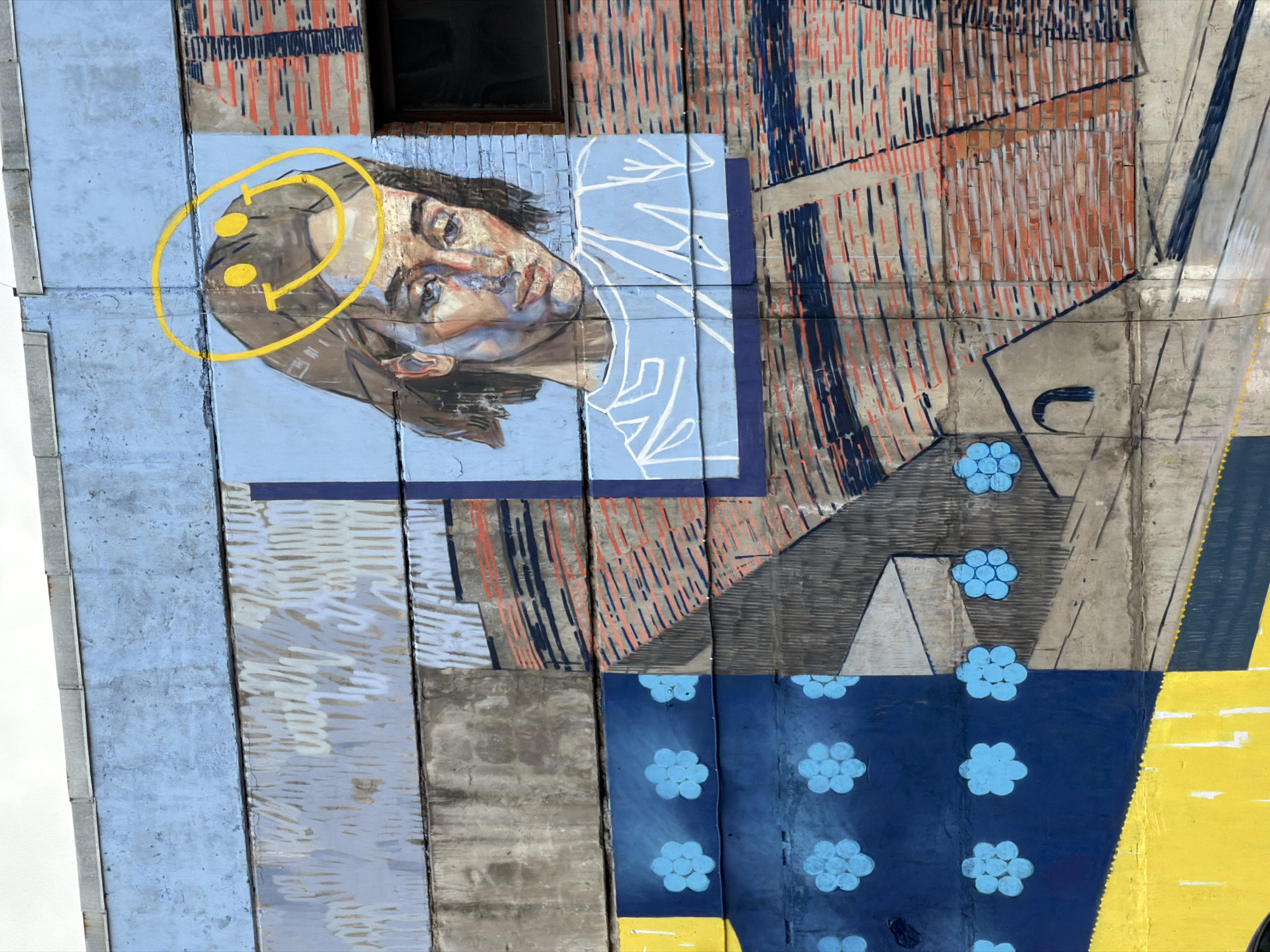 This is a photograph of a mural painted on the side of a building. The mural is composed of several distinct sections. The most prominent element is a portrait of a person with a yellow circle around the head and two yellow dots inside. The portrait is painted in blues and browns with white lines depicting clothing. The remainder of the mural is an abstract design of concrete-looking sections painted in shades of blue, gray, orange, and yellow. The bottom of the mural is dark blue with flower designs in pale blue. The painting has been sectioned off by vertical lines as well. The location of this image is in the city of Saint Petersburg, Russia.