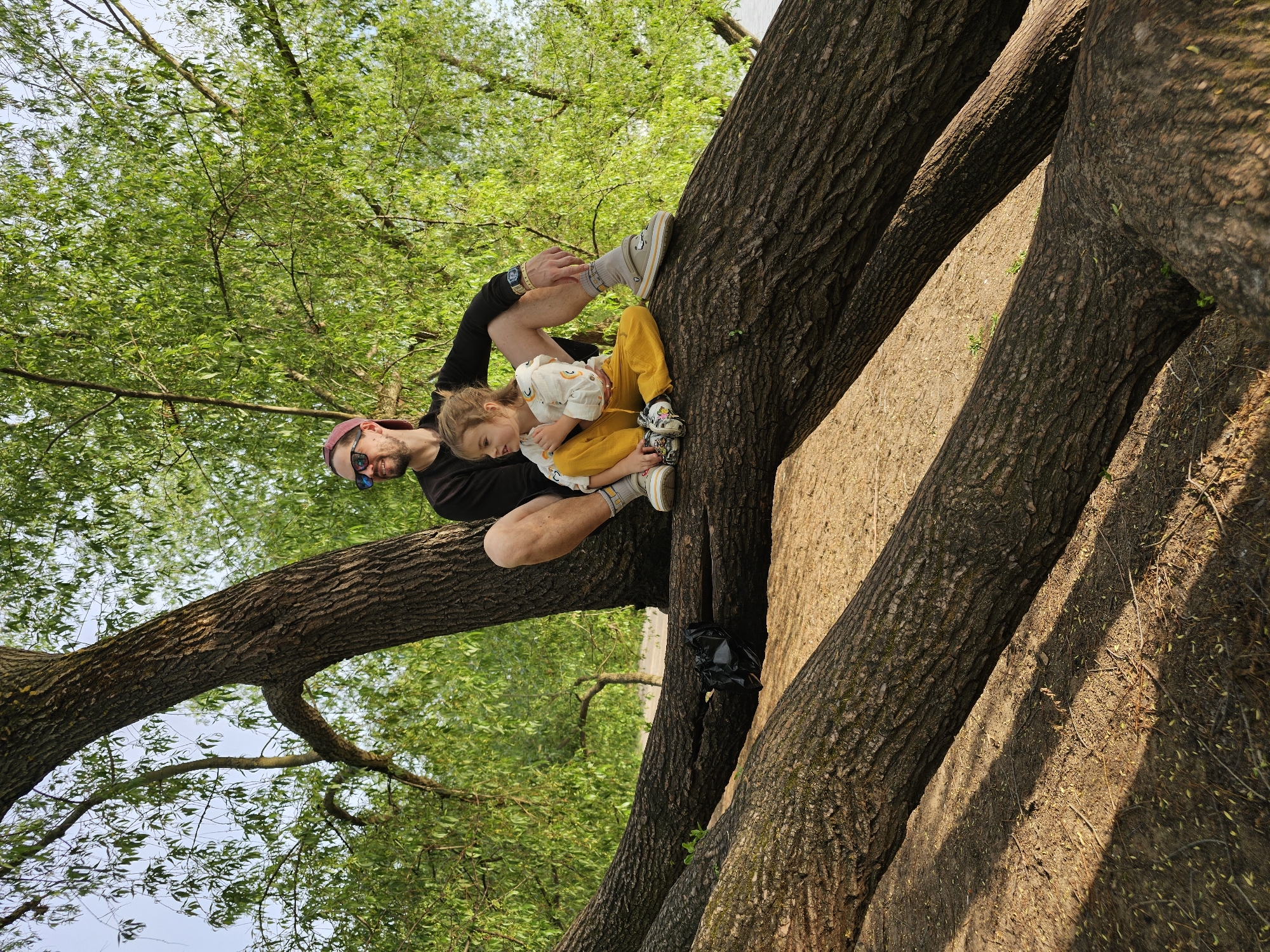A man and a young child are positioned in a tree in Khimki, Russia. The man, wearing a black t-shirt, a red baseball cap, and sunglasses, is seated on a large tree branch, holding the child in his lap. The child is wearing a white shirt with rainbows, yellow pants, and Crocs shoes. The branches are thick and textured, with leaves visible. The ground below the branches is covered in dirt and leaves. A black plastic bag is seen nestled between the tree branches. The sky is visible through the leaves in the upper portion of the image.