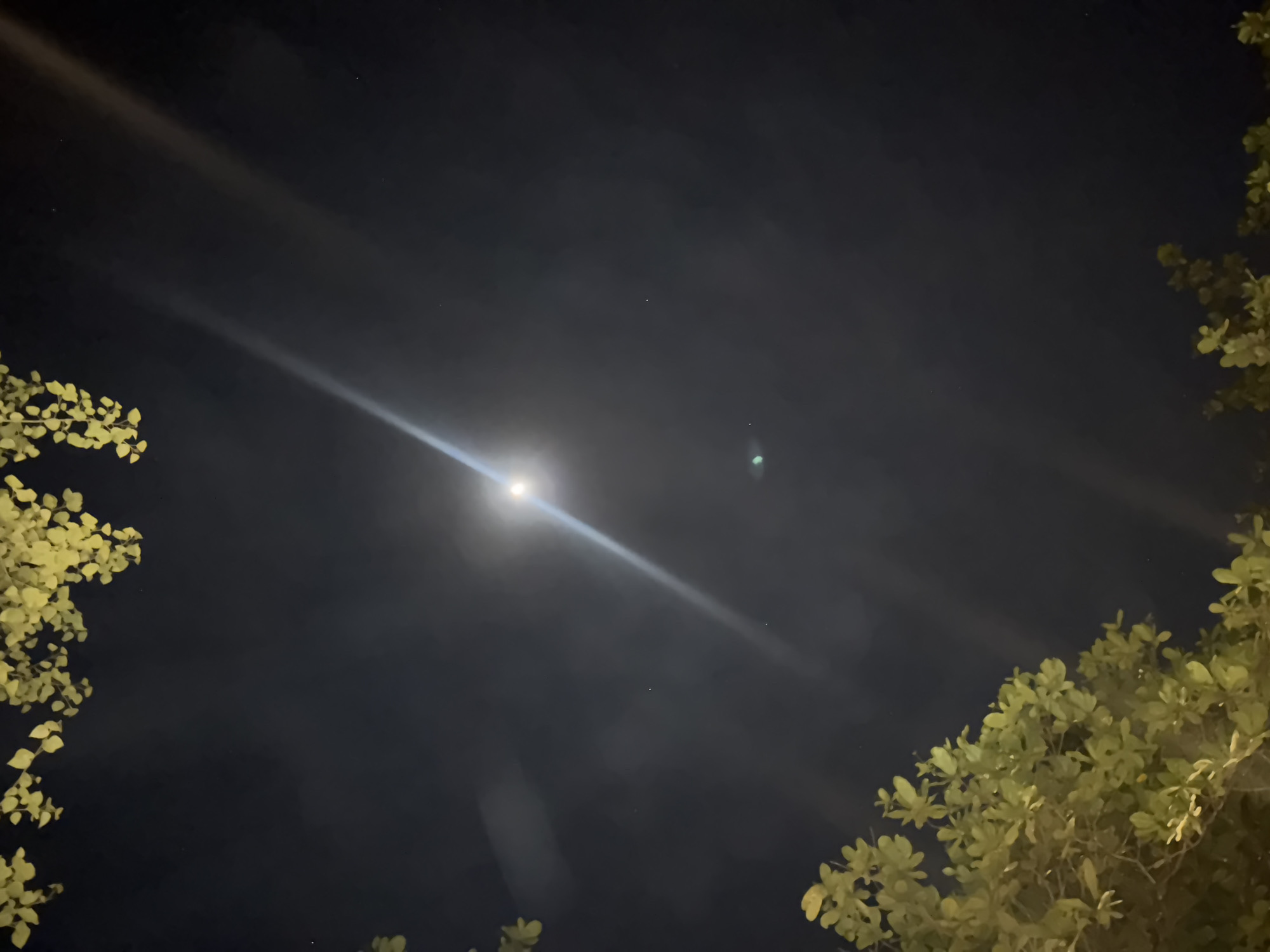 The image shows a night sky with a bright light source, likely the moon, emitting a strong beam. The sky is dark with some visible clouds. Green foliage is present on the left and right sides of the frame. The location appears to be Kuta Selatan, Indonesia. There are no people depicted in the image. The scene captures a nighttime outdoor environment with natural elements.